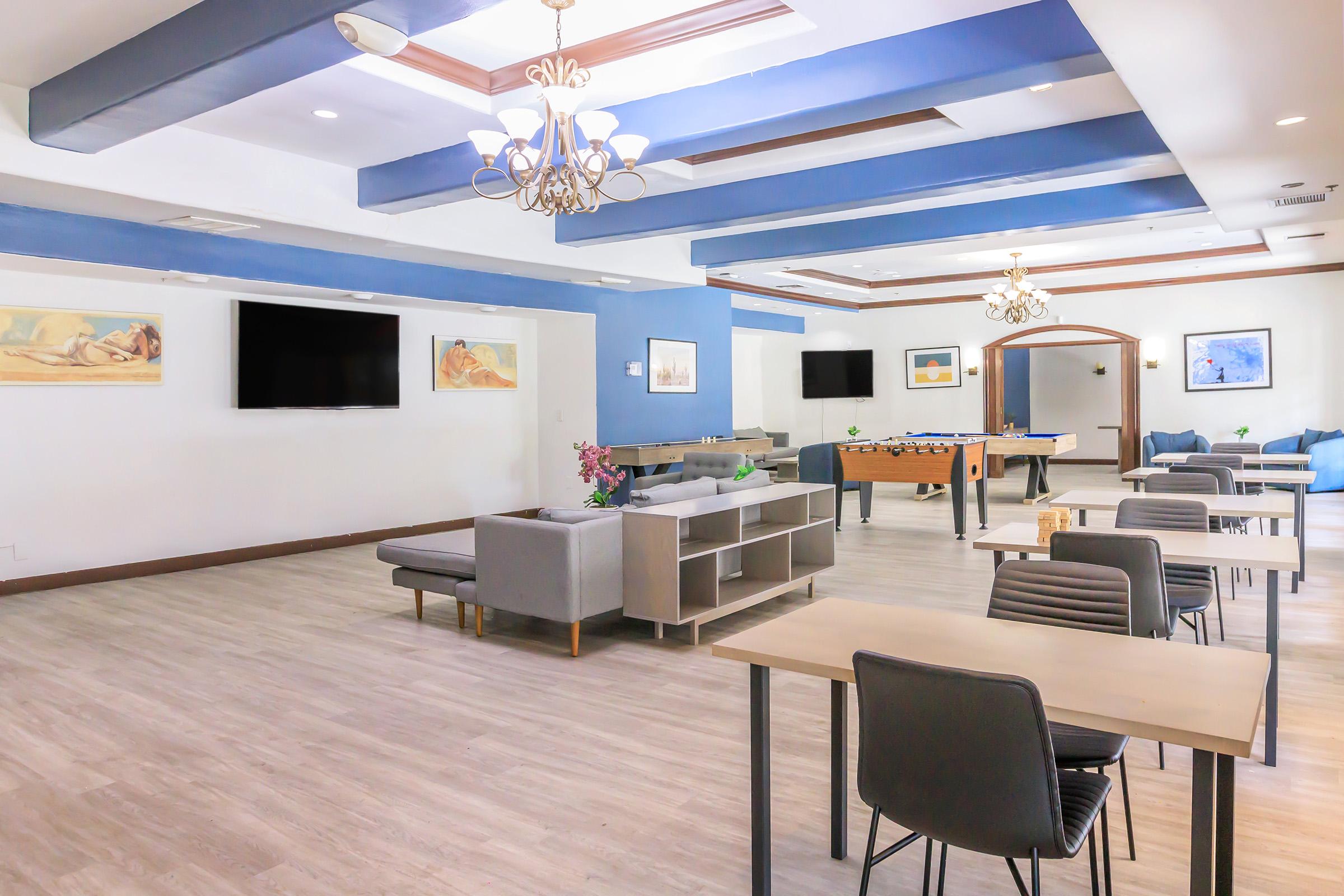 A modern lounge area featuring gray sofas and tables, with blue accent walls. Two large flat-screen TVs are mounted on the walls. A foosball table is visible in the center, along with decorative art pieces. Natural light illuminates the space, creating a welcoming atmosphere for relaxation and socializing.