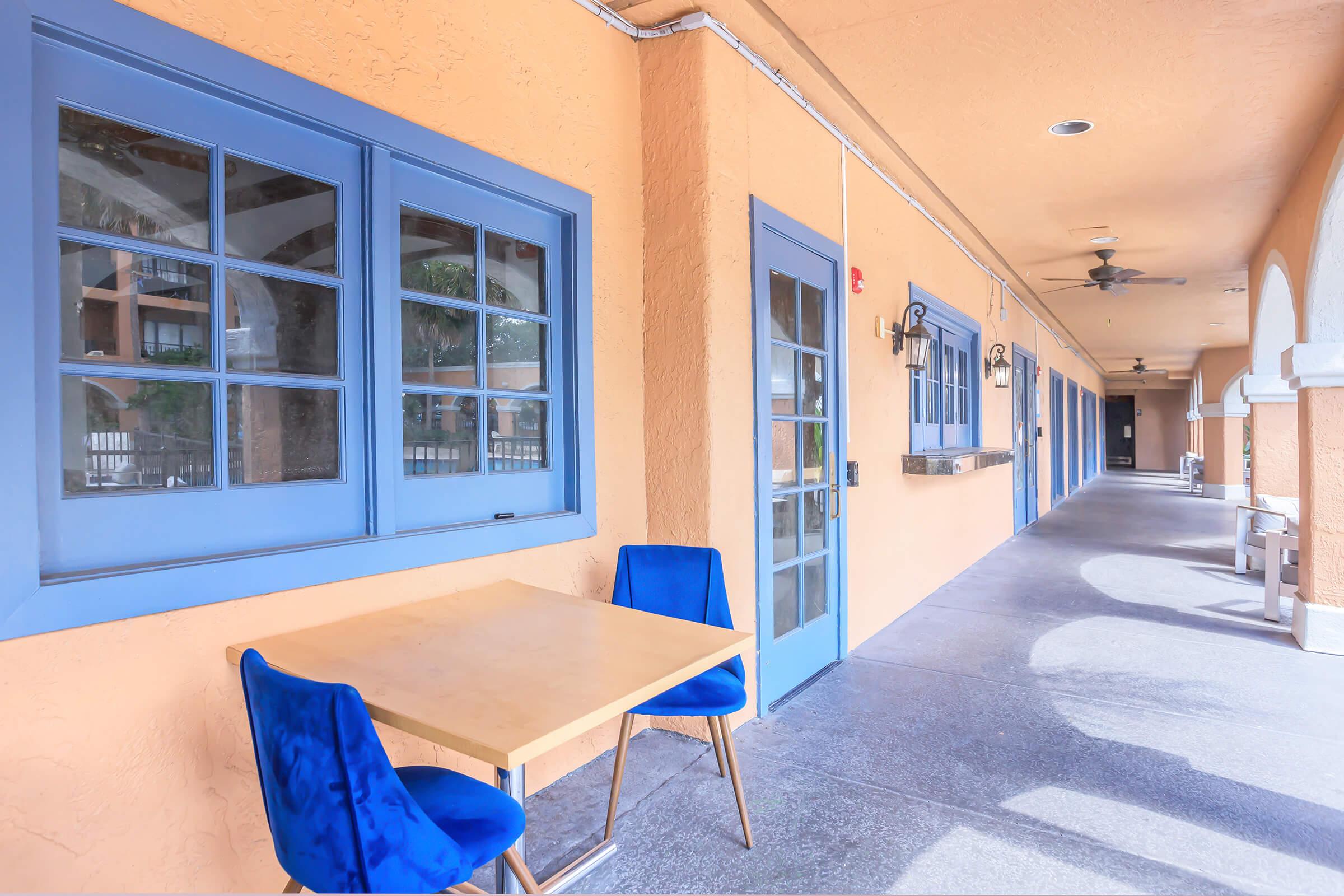 A hallway of a building featuring an orange wall with blue-framed windows and doors. There is a small wooden table with blue velvet chairs set outside. Sunlight casts shadows on the gray pavement, creating a bright and inviting atmosphere.