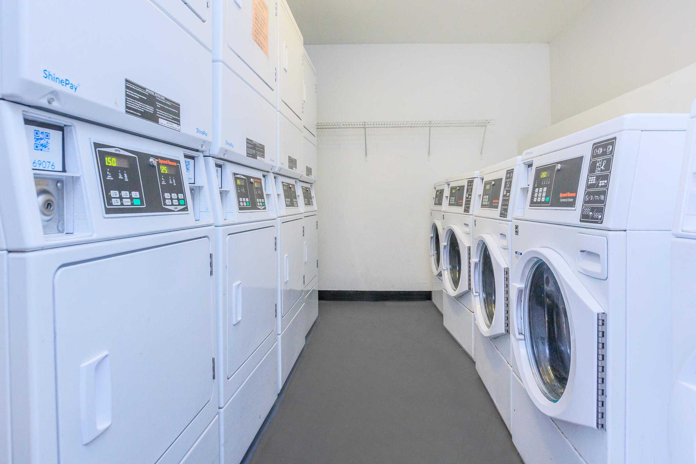A row of white laundry machines, including washers and dryers, in a clean, well-lit laundry room. The machines have digital displays and are lined up against a wall, with a simple, uncluttered floor and a shelf above.