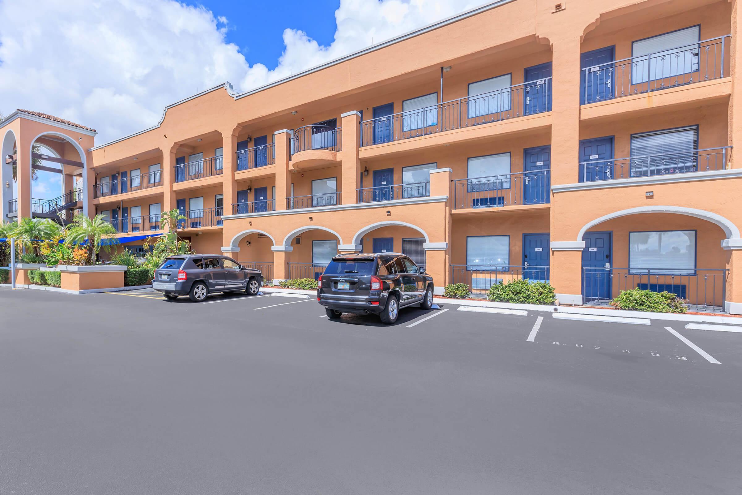 Two parked cars in front of a three-story orange building with multiple balconies and blue doors. The building features architectural arches and is surrounded by greenery under a partly cloudy sky. The parking lot is marked with white lines.