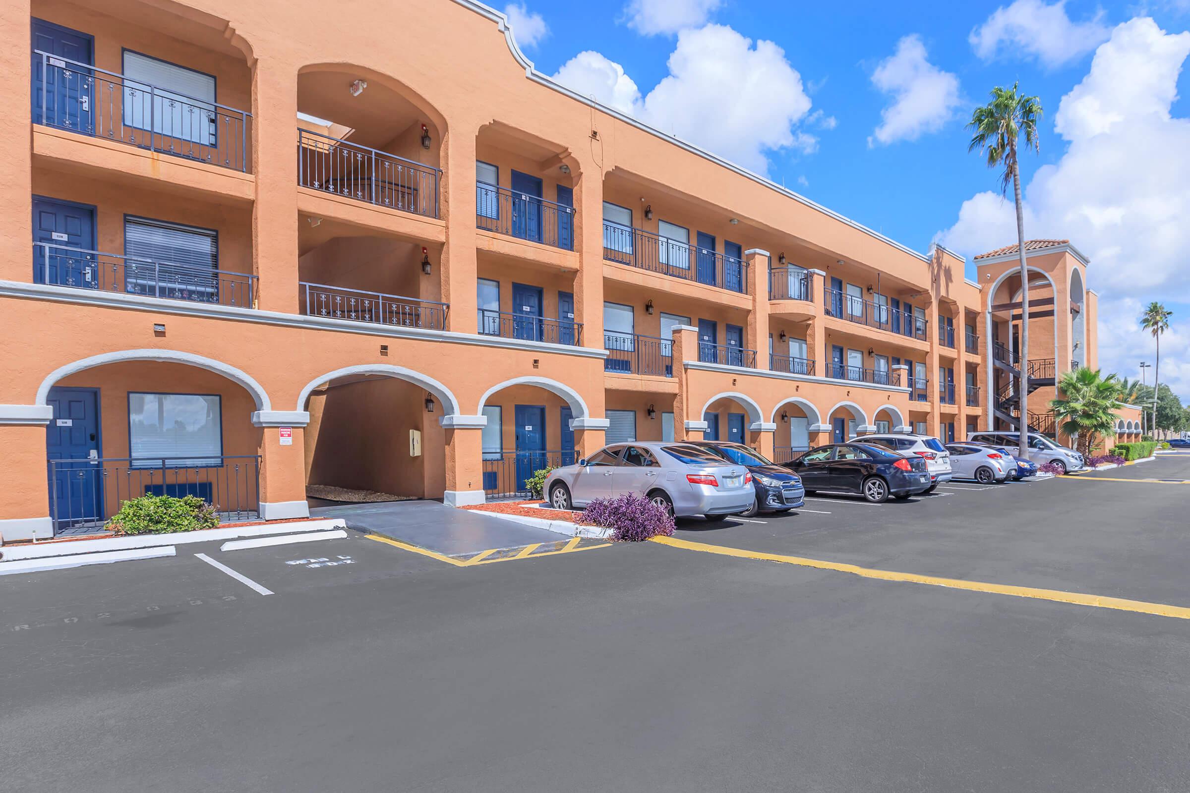 Exterior view of a multi-story orange building with balconies, featuring a parking lot filled with several cars. Palm trees and green landscaping enhance the scene, under a bright blue sky with fluffy white clouds. The structure has an arched architecture around the entrance and balconies.