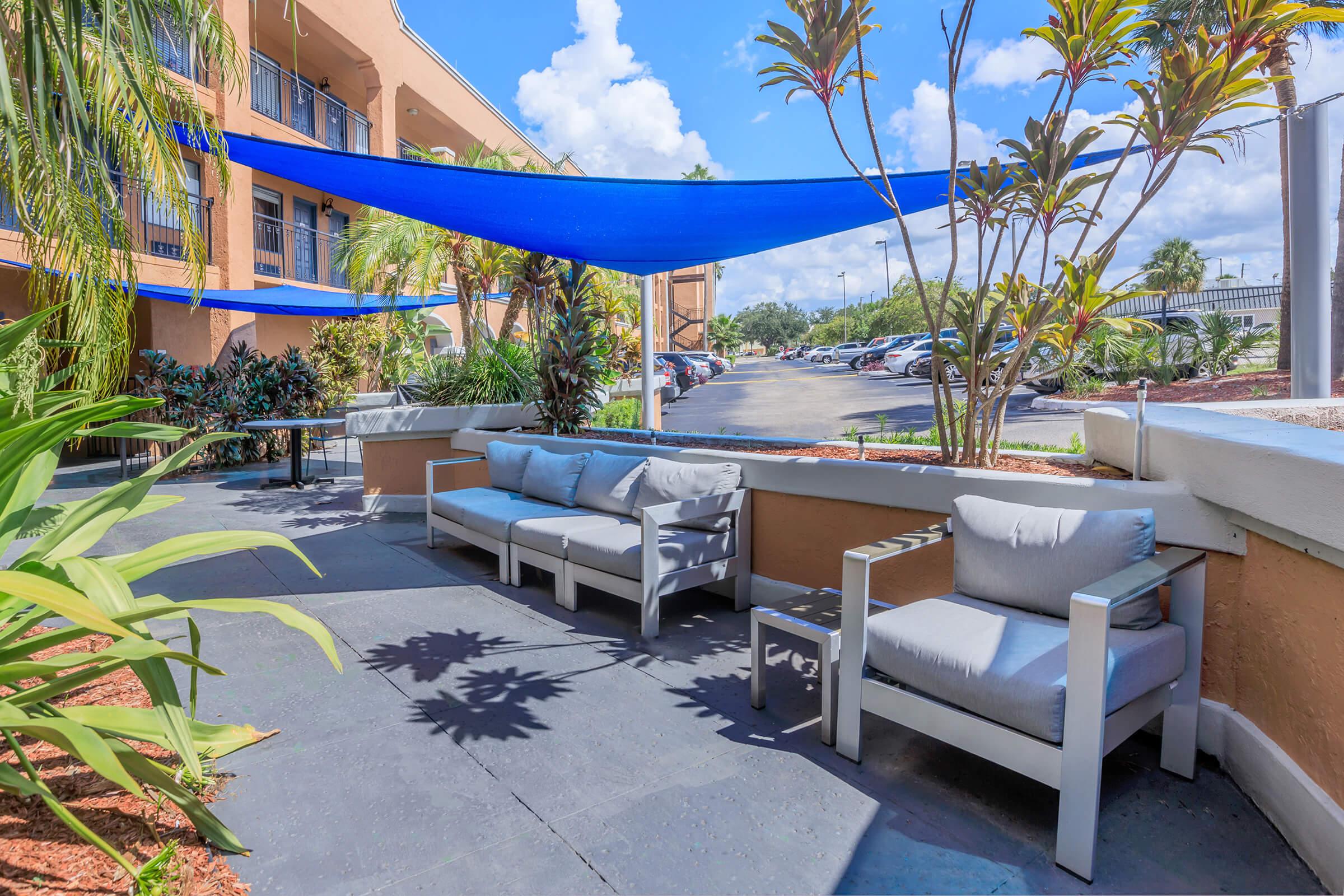 A cozy outdoor seating area featuring light gray sofas with white frames under a blue shade sail. Surrounding greenery adds a lush, tropical feel. In the background, a parking lot is visible, with cars parked under a bright blue sky and scattered clouds.
