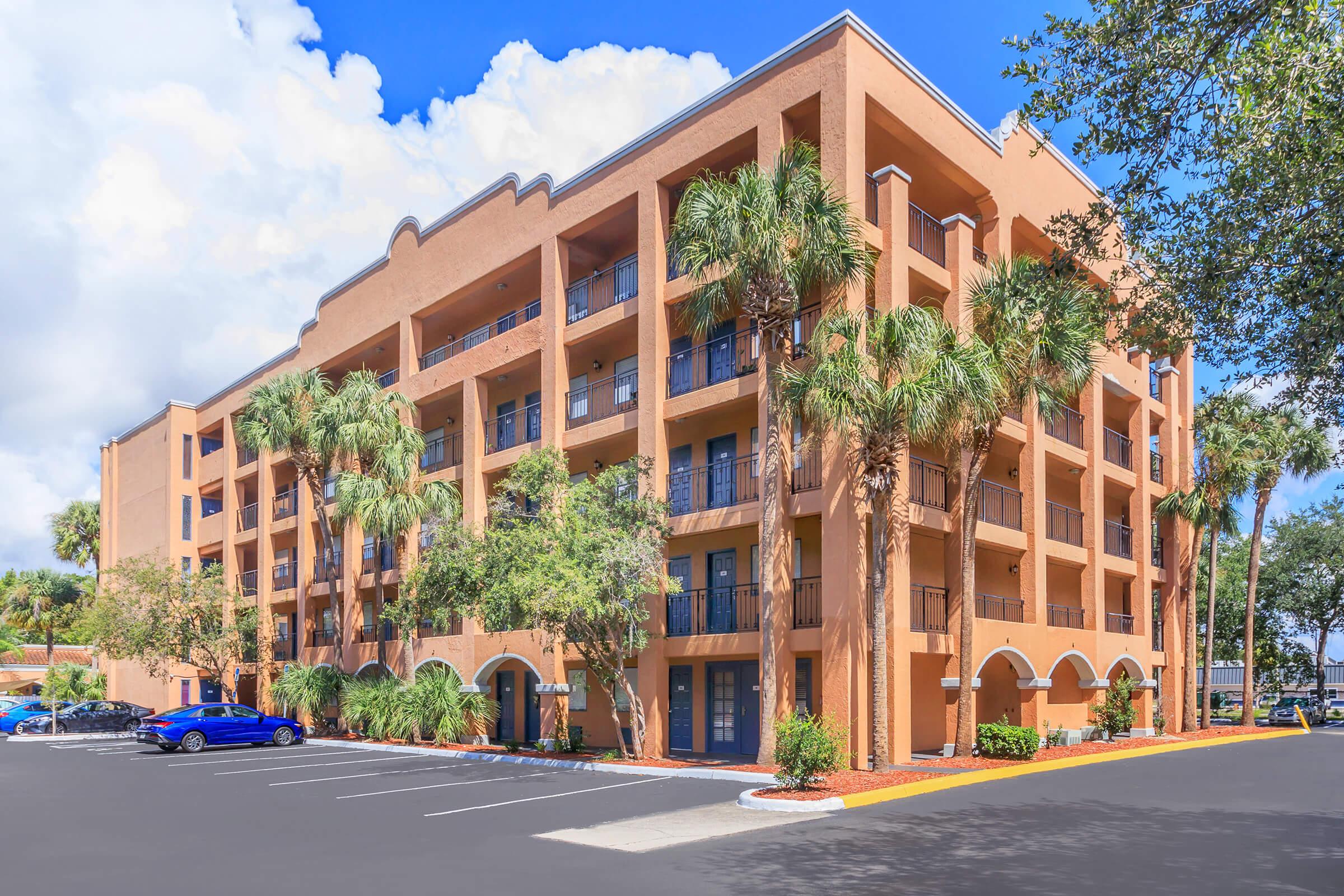 A modern, three-story building with a warm orange facade, featuring balconies and large windows. Palm trees and greenery surround the structure, which is situated in a parking lot under a bright blue sky with fluffy clouds.