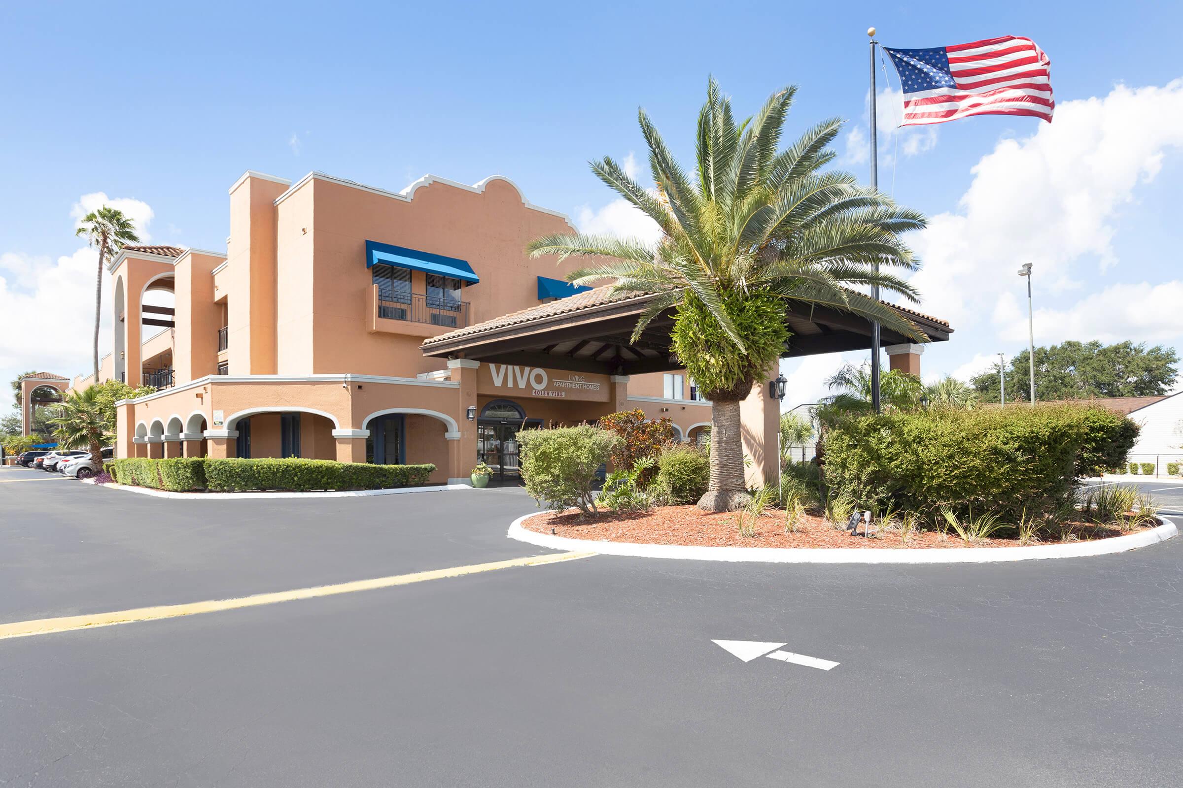 The image shows the exterior of a hotel with a prominent entrance featuring tropical landscaping and a flagpole displaying the American flag. The building has a warm orange color with blue awnings and a welcoming drive-up area. Clear blue skies complement the scene, enhancing the inviting atmosphere.