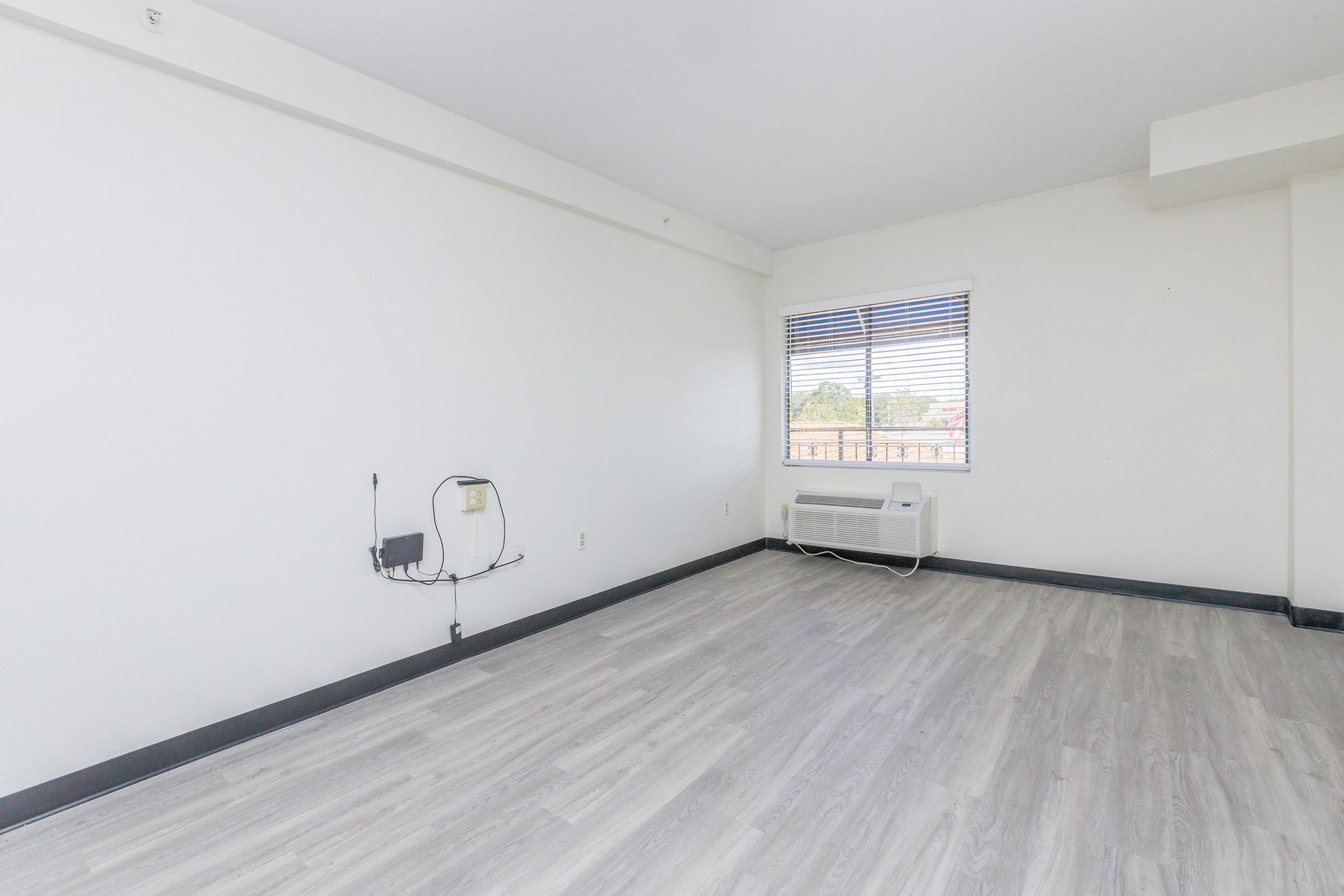 Empty room with light gray wooden flooring, white walls, and a window with blinds. An air conditioning unit is partially visible on the floor. There are no furnishings or decorations present, giving the space a minimalistic appearance.