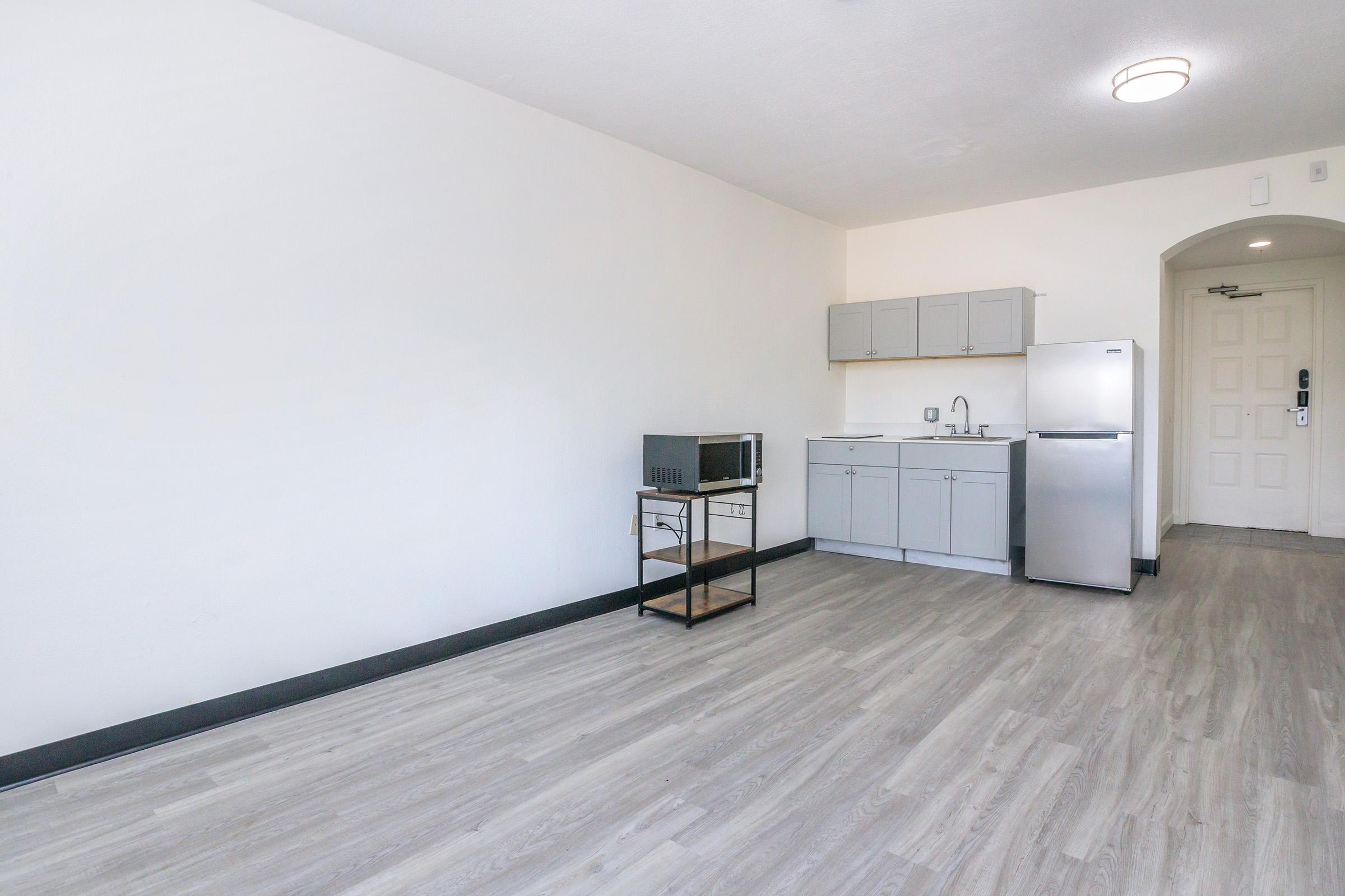 A spacious, minimalistic interior of a room featuring a small kitchenette with gray cabinets, a sink, a microwave, and a refrigerator. The flooring is light-colored, and there are no furnishings in the room. A door is visible in the background, leading to an entrance. The walls are painted white, enhancing the open feel.