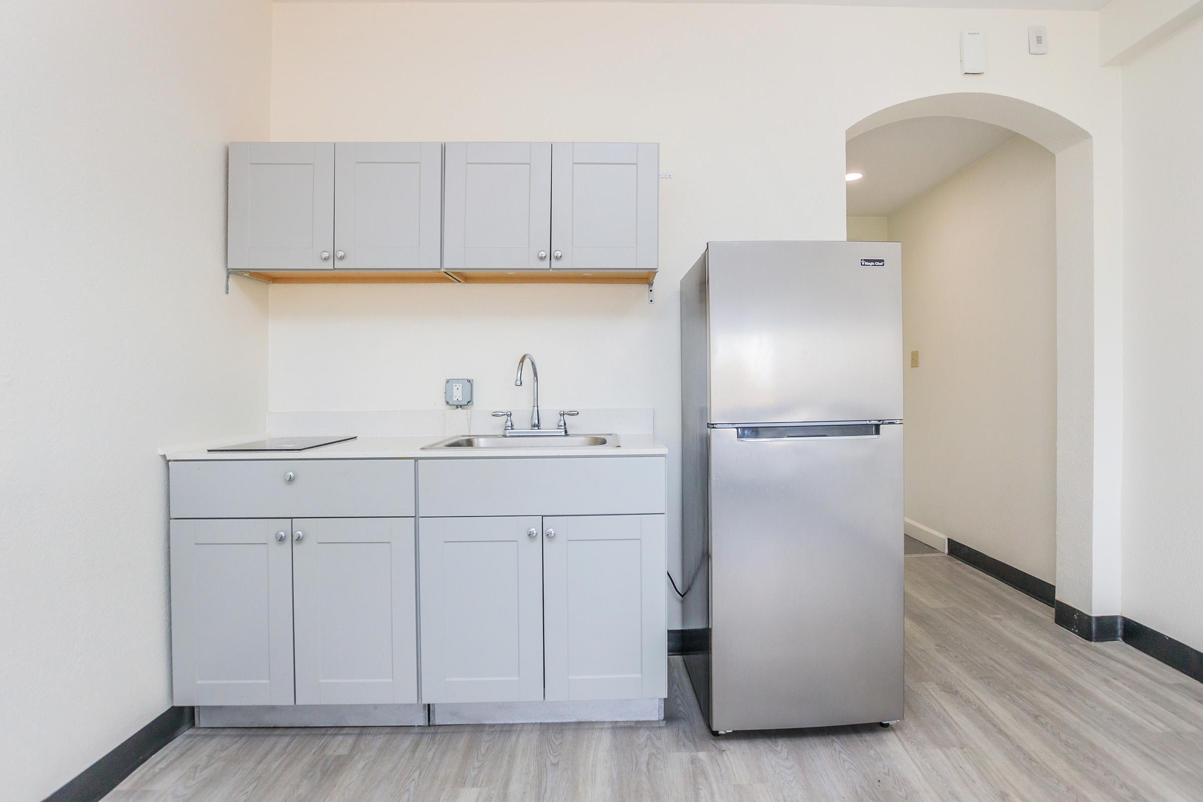 A clean, modern kitchenette featuring gray cabinetry, a stainless steel refrigerator, and a sink. The space has light-colored walls and a gray wood-style floor, with an open layout and a doorway leading to another area.