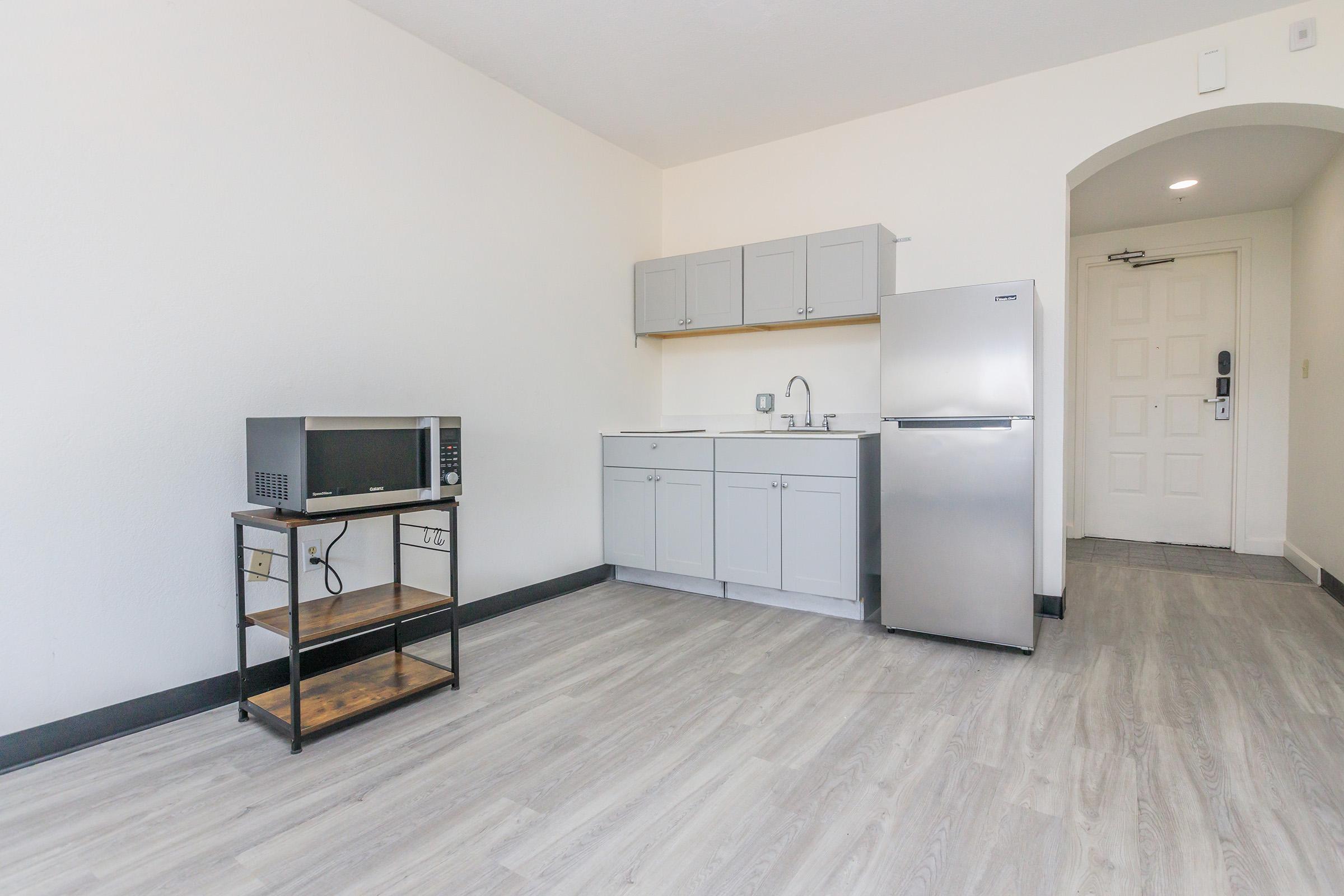 A small, modern kitchenette featuring a stainless steel refrigerator, a microwave on a wooden shelf, and gray cabinets. The room has light-colored walls and wooden flooring, with a simple door at the back, creating a minimalistic and open feeling. Natural light brightens the space.