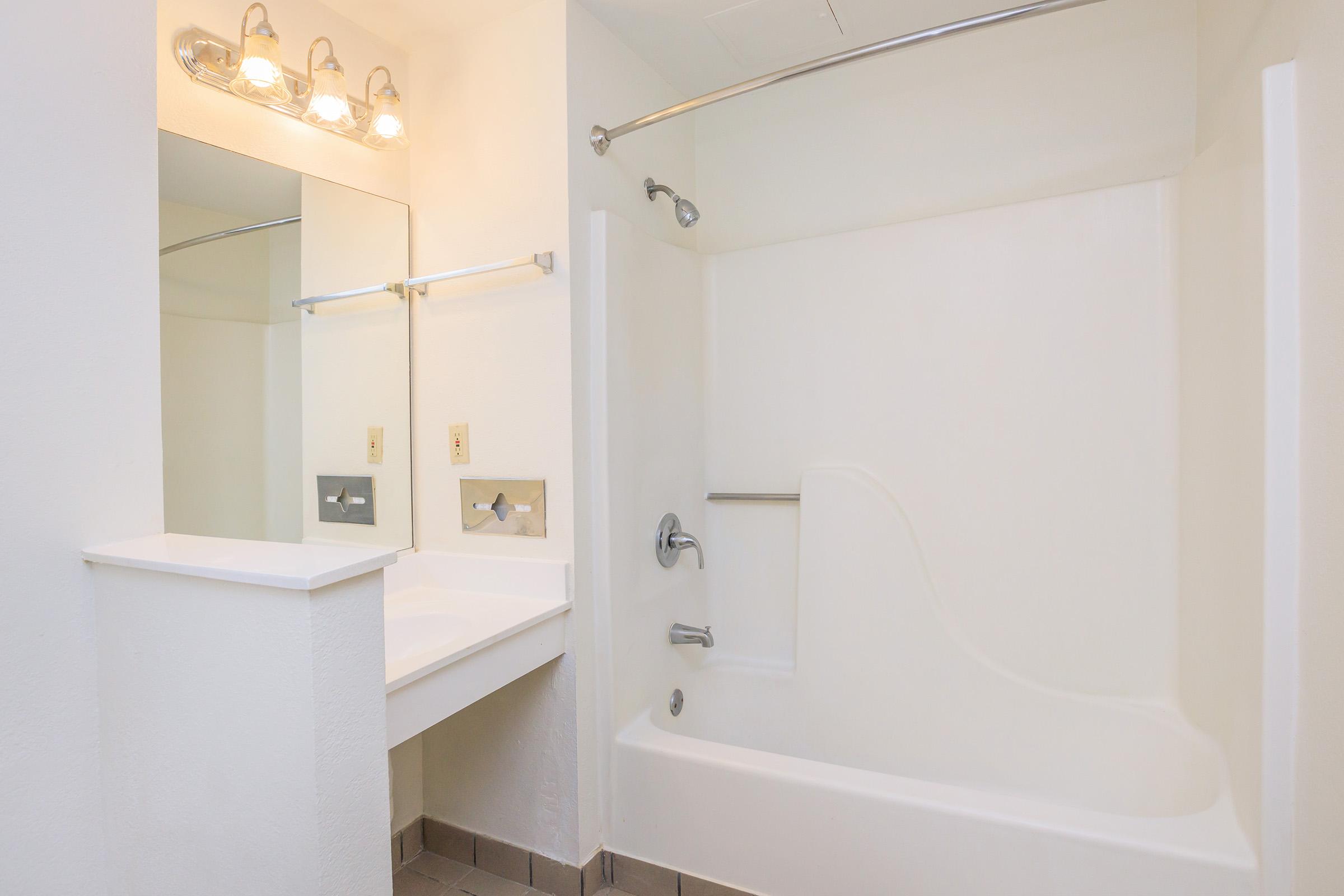 A clean, modern bathroom featuring a white bathtub with a shower, a large mirror above a sink with a countertop, and bright lighting fixtures. The walls are painted white, with tiled flooring adding a touch of texture.