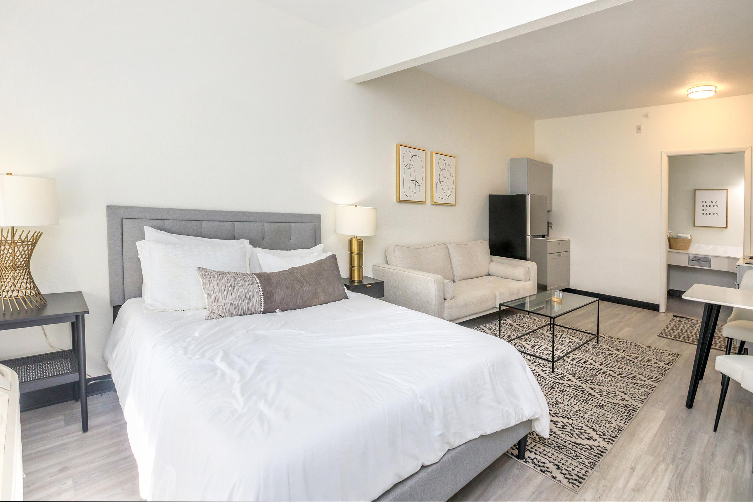 A well-lit studio apartment featuring a gray upholstered bed with white bedding, a modern gray sofa, a glass coffee table, and two lamps on nightstands. The kitchenette is partially visible, with a refrigerator and a small dining area in the background. The decor is minimal and contemporary.