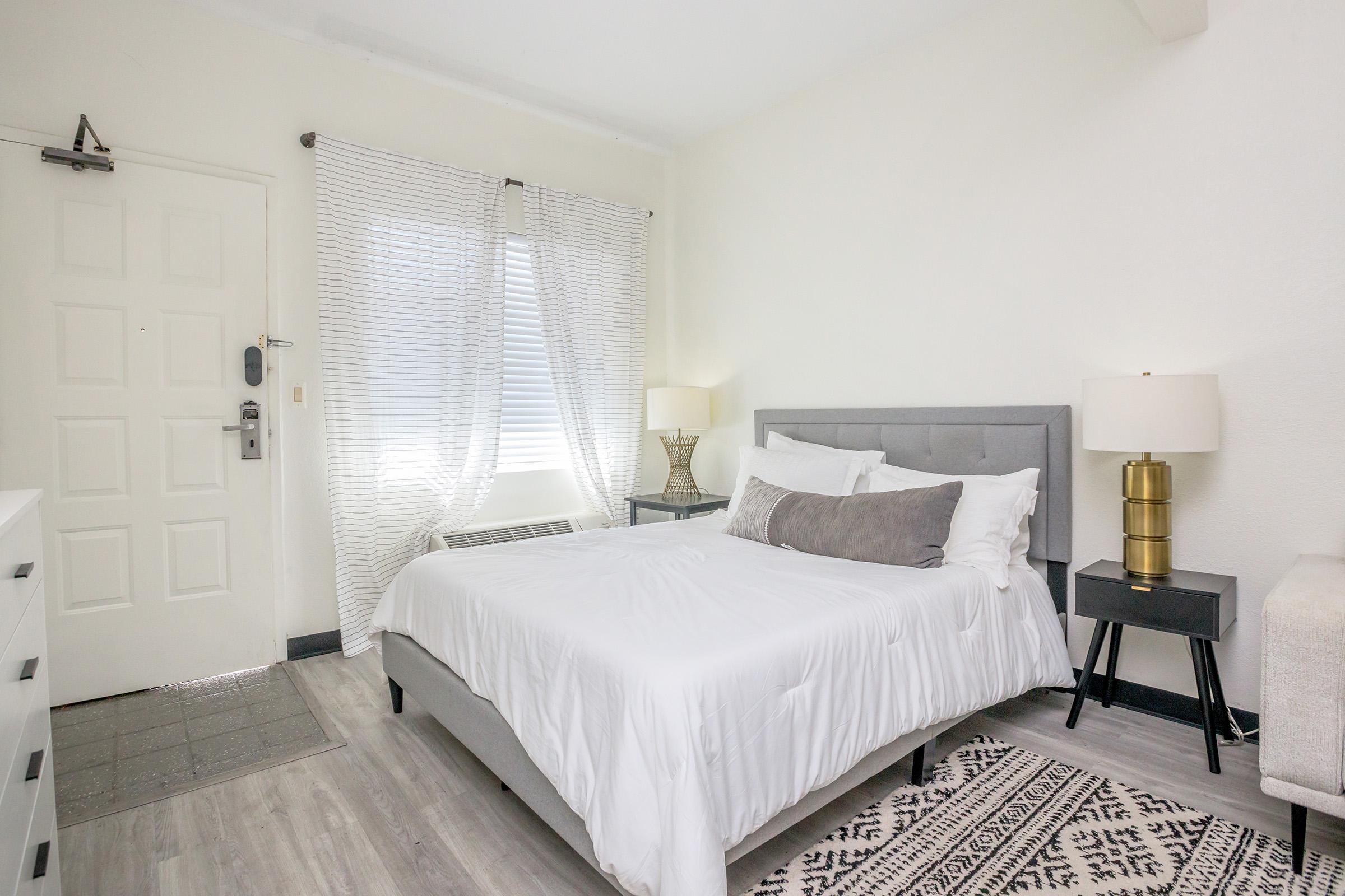A bright, modern bedroom featuring a queen-sized bed with white bedding, a gray upholstered headboard, and decorative pillows. There are two bedside tables with stylish lamps, a window with sheer curtains, and a minimalist decor style, including a small dresser and a patterned rug on the floor.