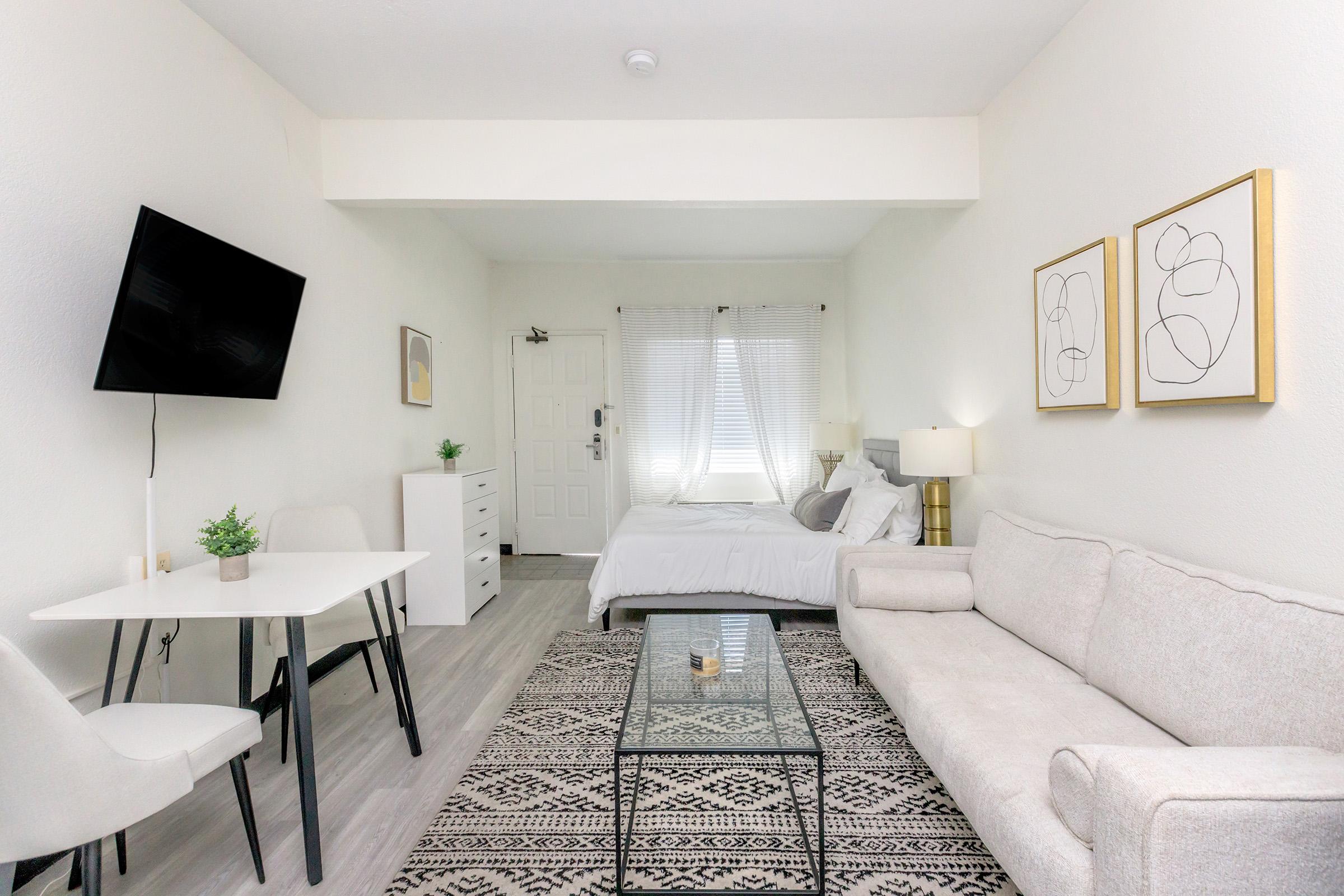 A modern, minimalist living space featuring a small dining table with two chairs, a light-colored sofa, and a glass coffee table. A wall-mounted TV is positioned opposite a bed with white bedding. Natural light filters through two windows, and abstract art is framed on the wall.