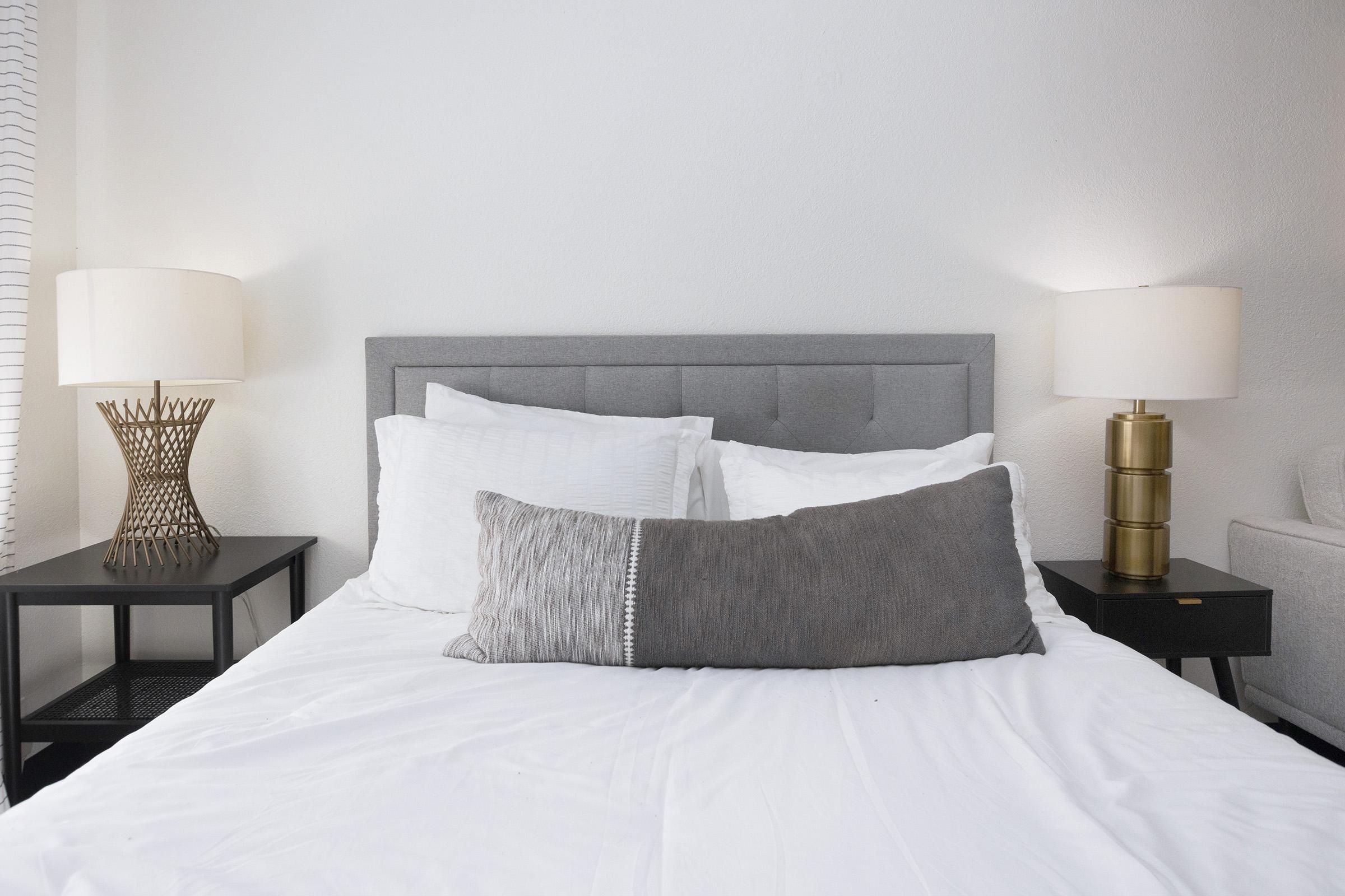 A neatly made bed with white linens and a gray tufted headboard. There is a long gray decorative pillow in the center. On either side of the bed, there are modern nightstands, one with a decorative lamp featuring a geometric design and the other with a cylindrical lamp, both providing soft lighting.