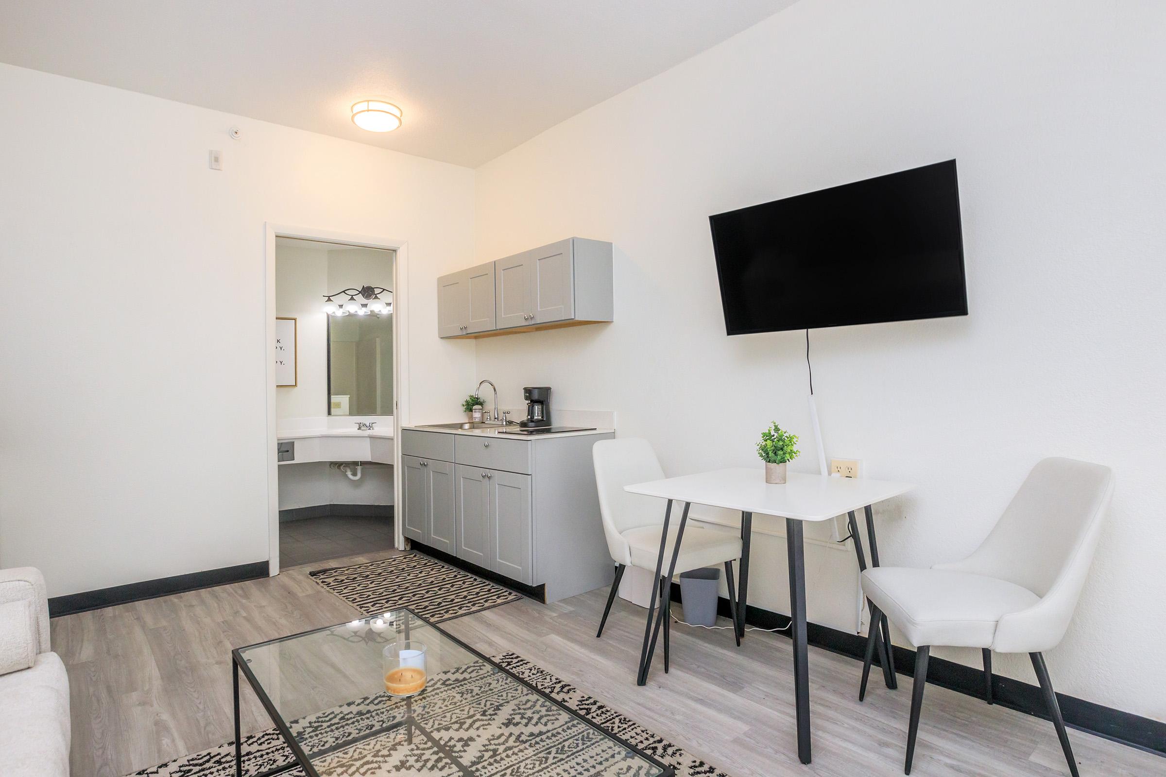 A modern, minimalist studio apartment featuring a small kitchenette with gray cabinets, a coffee maker, and a flat-screen TV mounted on the wall. There is a small dining table with two chairs and a cozy seating area with a light-colored couch. A bathroom is visible in the background.