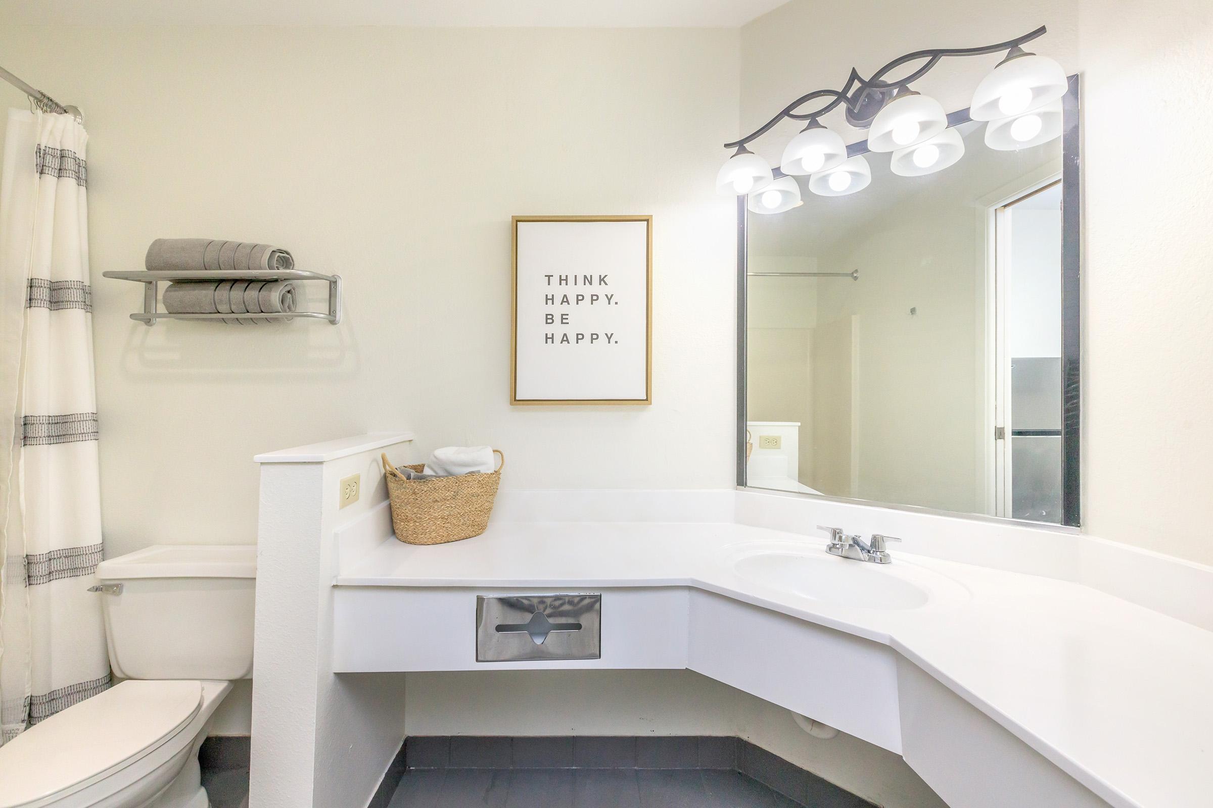 A bright bathroom featuring a white countertop with a sink, a mirror above, and a wall light fixture with multiple bulbs. There's a framed motivational quote that reads "THINK HAPPY. BE HAPPY." A towel rack with neatly stacked towels, and a wicker basket sit beside the sink, adding to the clean and organized aesthetic.