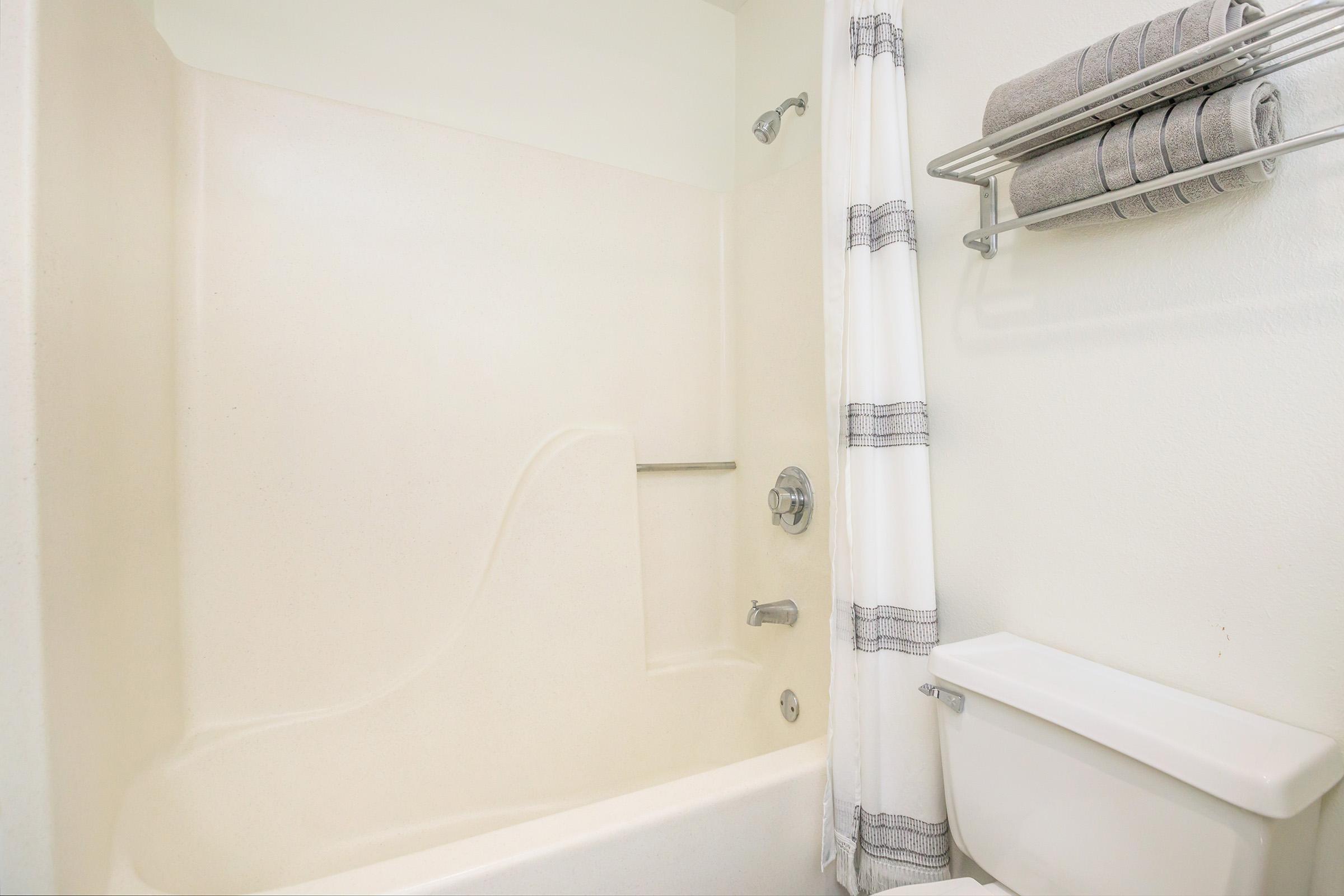 A clean bathroom featuring a simple white bathtub with a shower, a silver towel rack holding neatly rolled gray towels, and a white toilet beside it. The walls are light-colored, enhancing the bright and spacious feel of the area.
