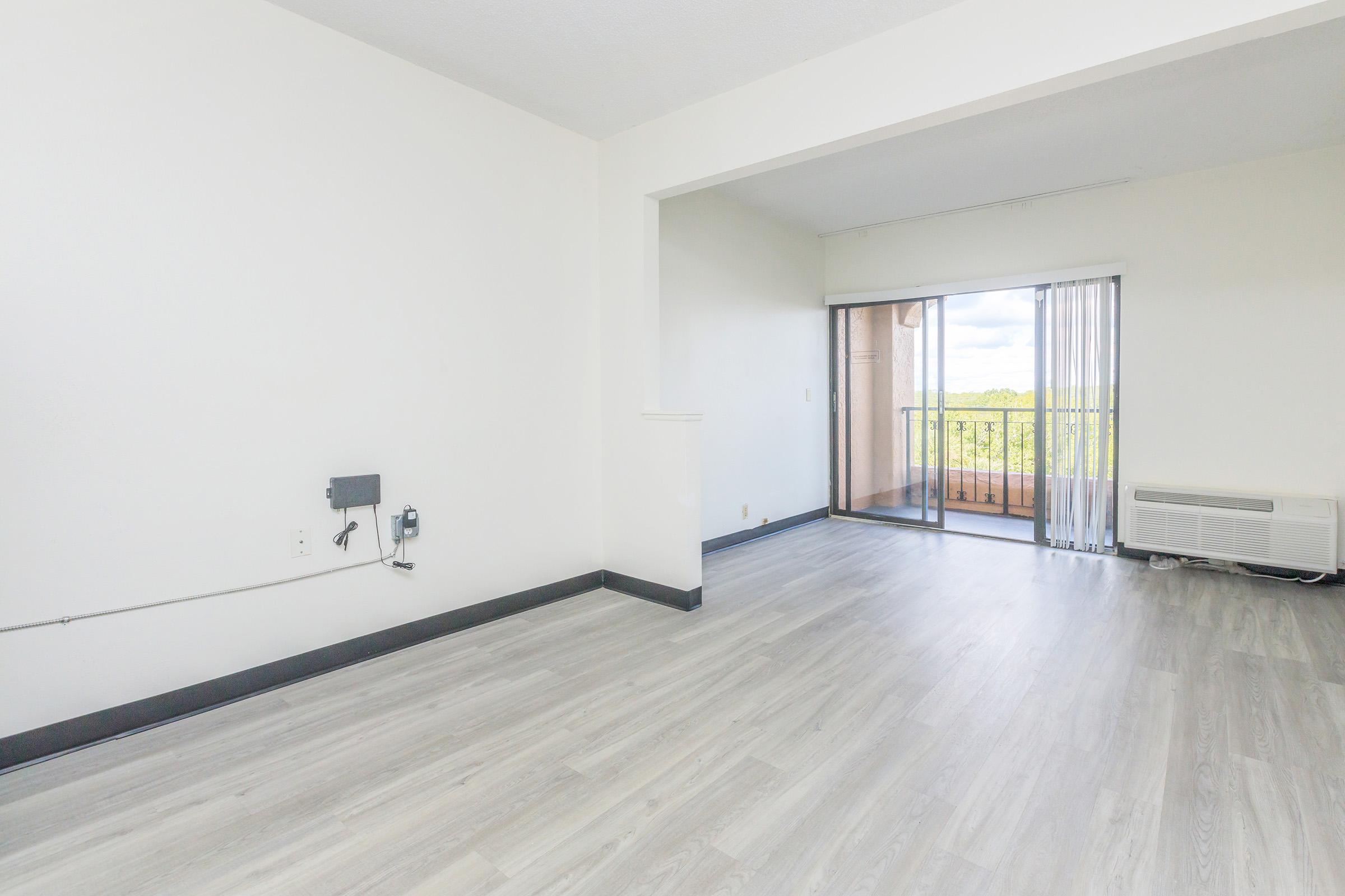 A spacious, empty living room with light-colored laminate flooring, white walls, and a sliding glass door leading to a balcony. An air conditioning unit and a wall outlet are visible on one side. The room is bright and airy, with natural light coming through the glass door.