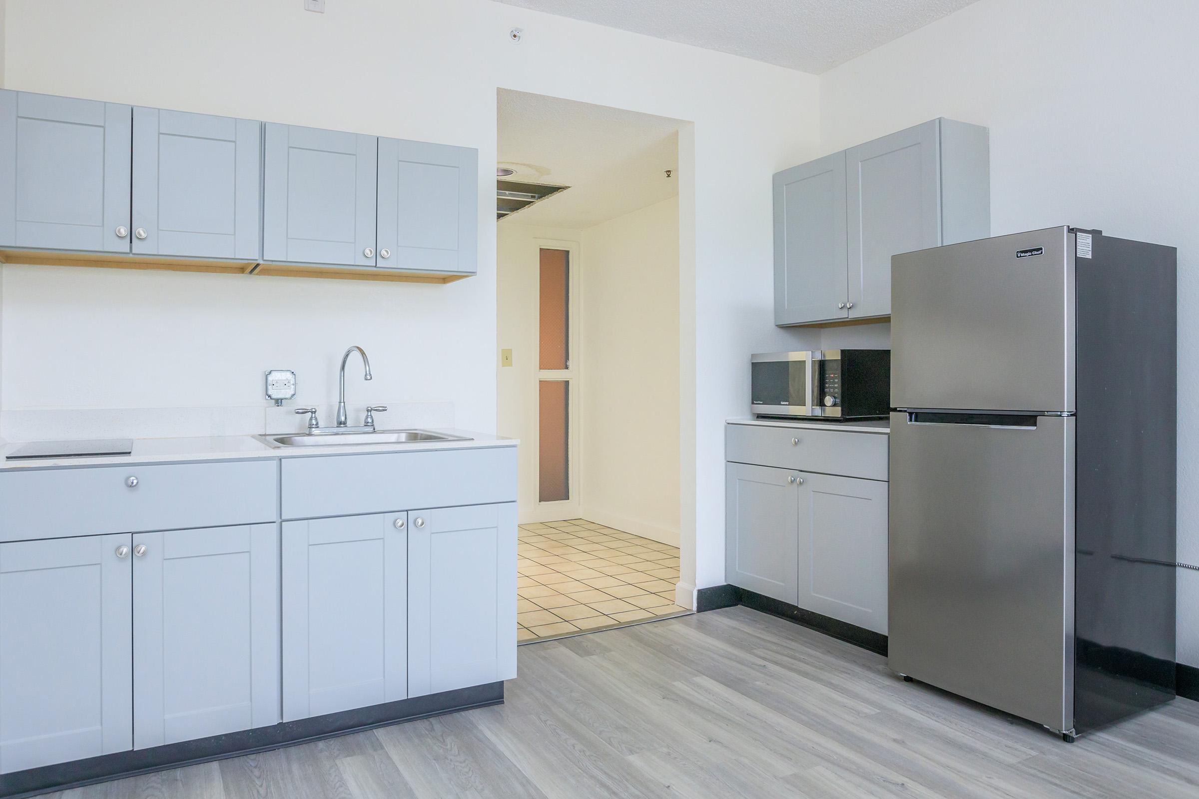 A modern, minimalist kitchen featuring light gray cabinetry, a stainless steel refrigerator, a microwave, and a sink. The kitchen has a wooden floor and opens into a hallway with beige tiles, creating a spacious and inviting atmosphere.