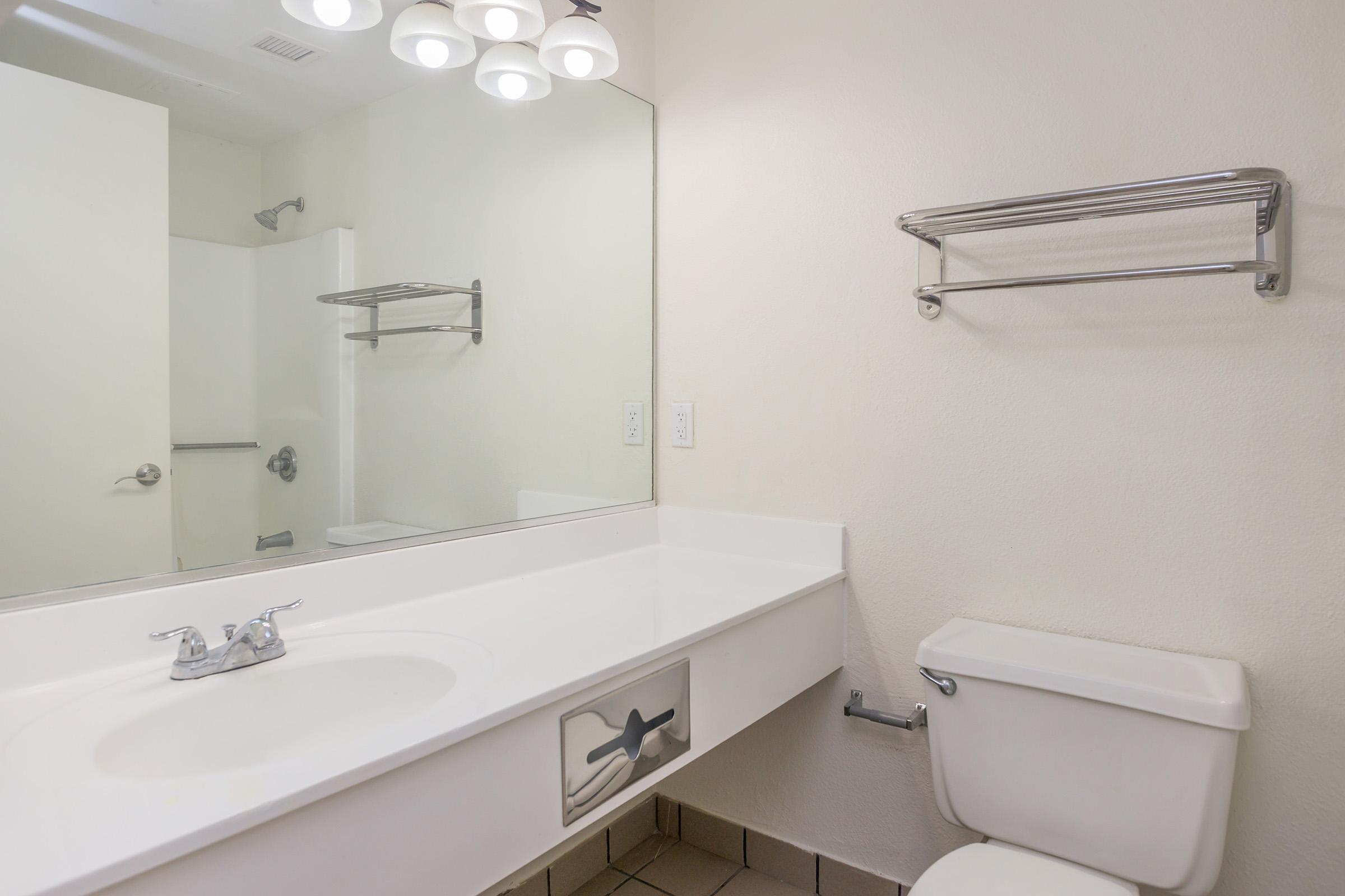 A clean bathroom featuring a white countertop with a sink, a large mirror above it, a toilet beside the sink, and a towel rack on the wall. The walls are light-colored, and there is a glimpse of a shower area in the background, providing a simple and functional design.