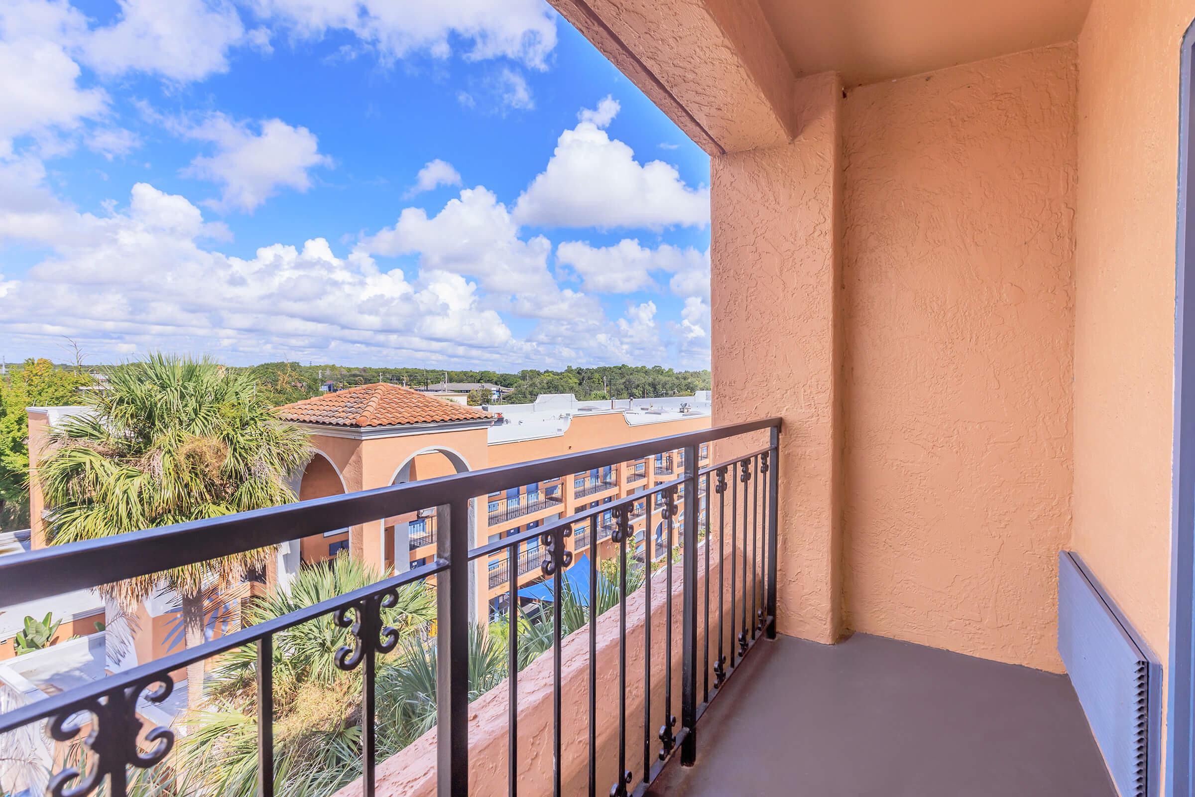A balcony view featuring a railing overlooking lush greenery and rooftops under a partly cloudy blue sky. The scene captures a serene outdoor space with vibrant colors and a peaceful atmosphere.