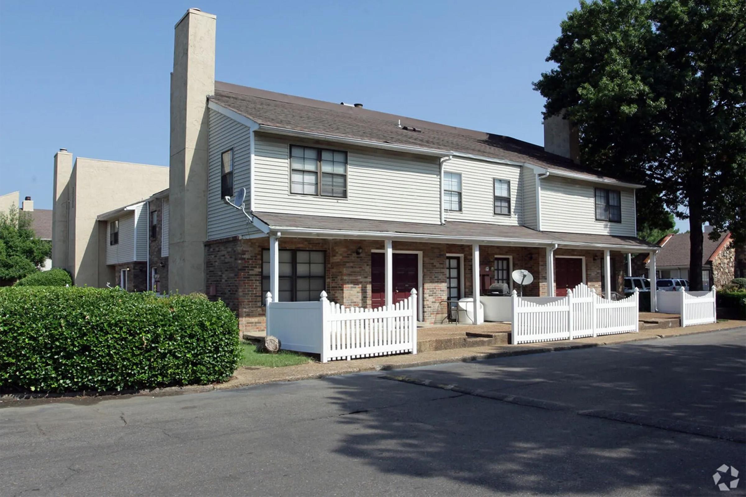 Two-story residential complex featuring several townhome-style units with light-colored siding and brick accents. Each unit has a small front porch and a white picket fence, with greenery and trees in the surroundings, and an asphalt driveway. The sky is clear and blue.