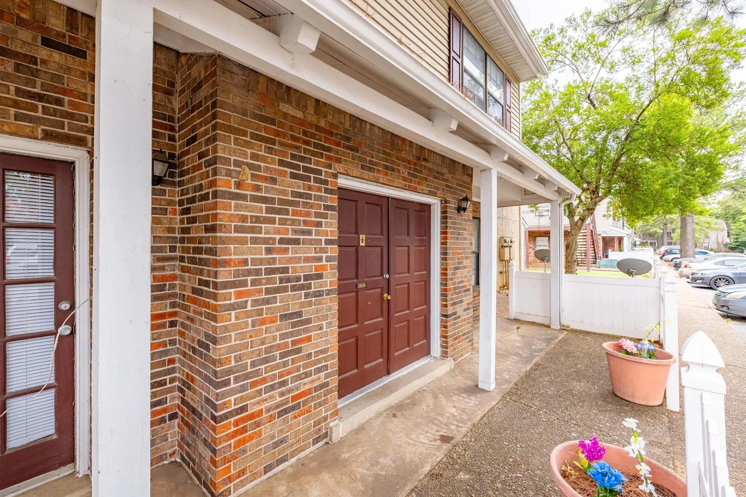 A brick exterior of a house showing a front door with a decorative design, flanked by two windows. Flower pots with colorful flowers are placed near the entrance. In the background, additional structures and tree foliage are visible, suggesting a residential neighborhood ambiance.