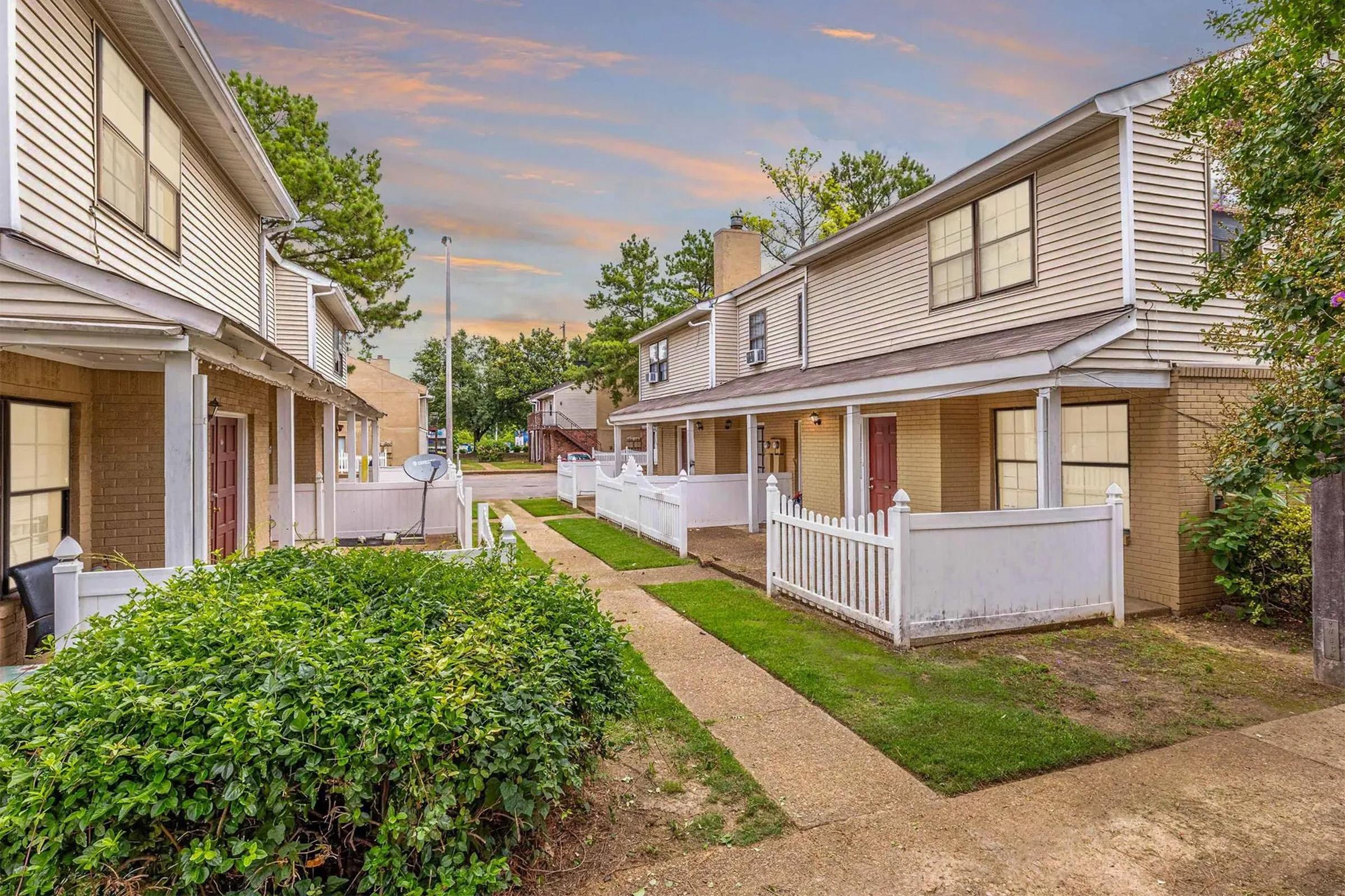 A view of a residential area featuring two-story townhouses with white picket fences. The pathway is lined with greenery, and trees can be seen in the background. The sky is painted with soft colors of sunset, adding a warm ambiance to the scene.