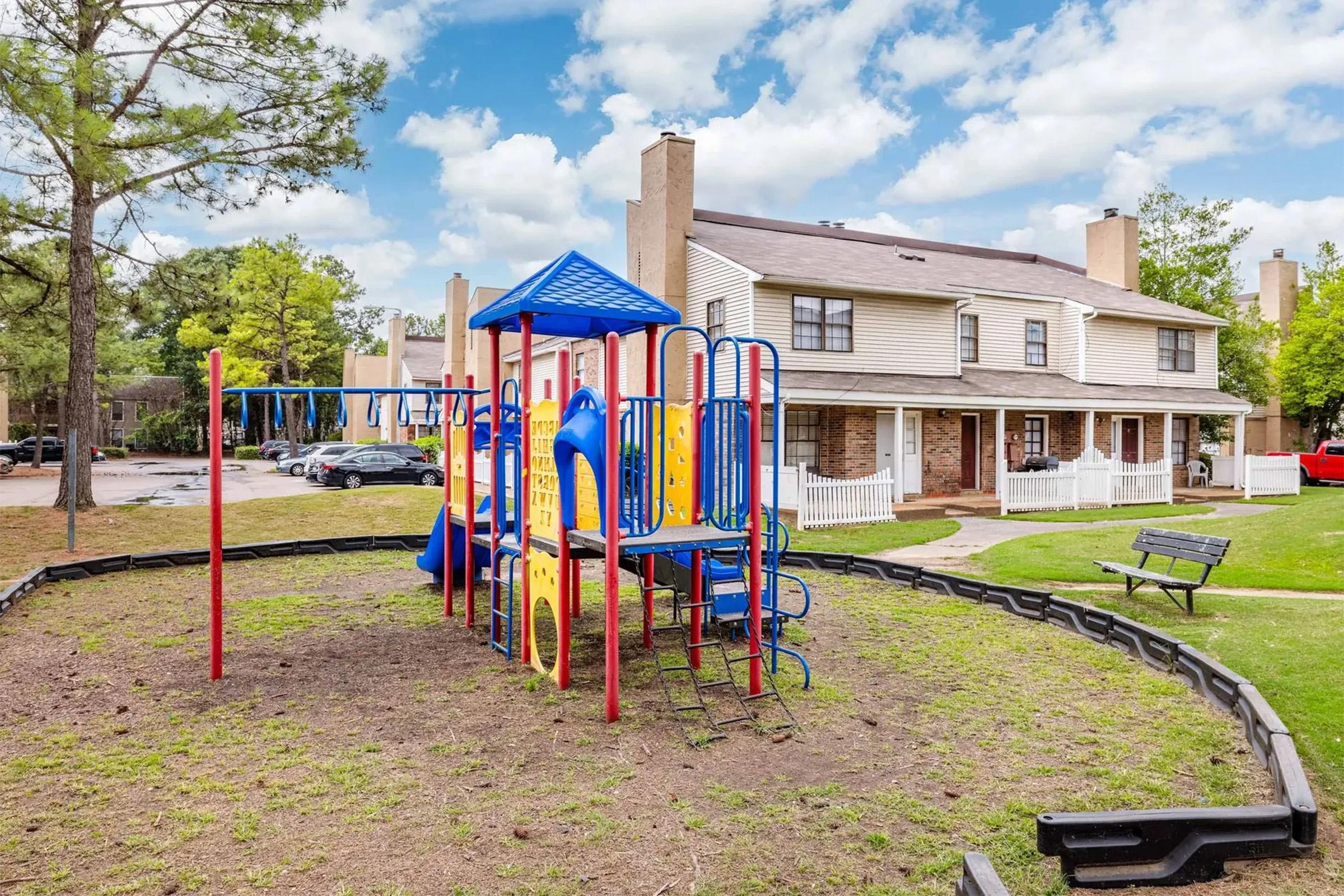 A colorful playground with slides and climbing structures is set on a grassy area next to a residential building. In the background, there are trees and a bench, with a few parked cars visible. The sky is partly cloudy, adding to the cheerful outdoor atmosphere.