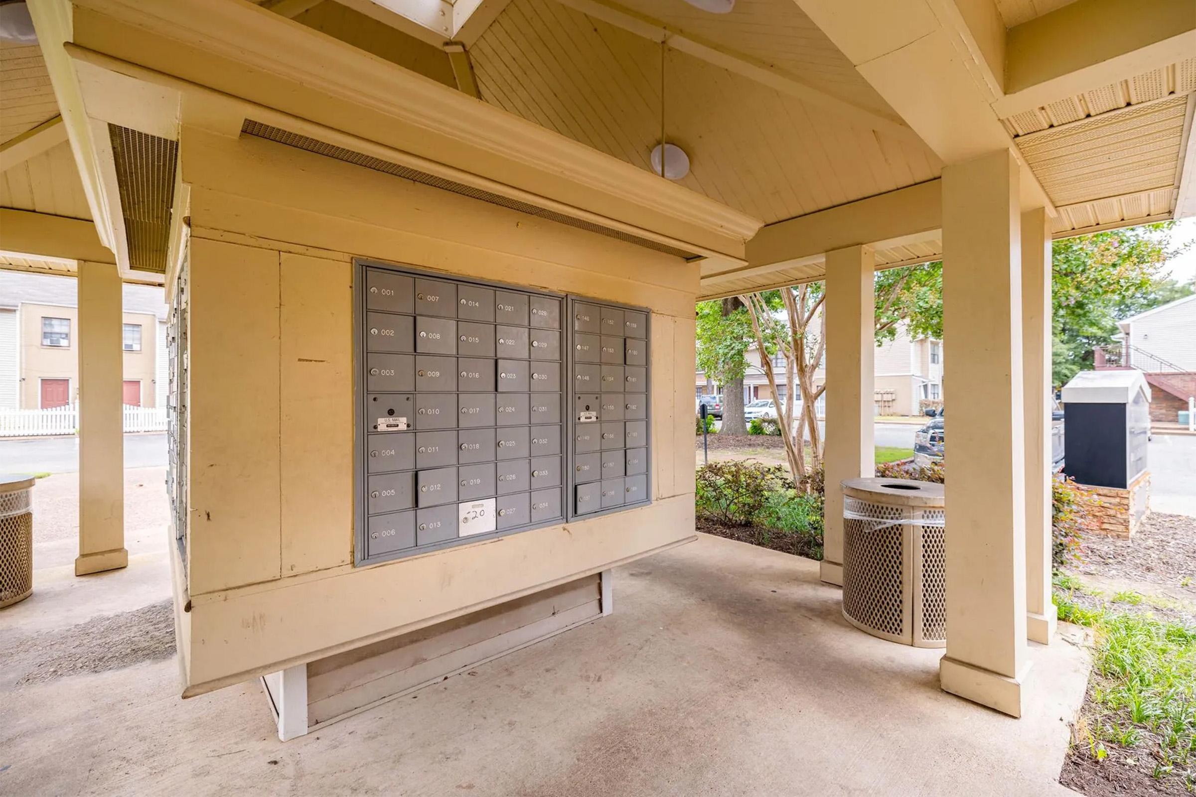 A row of gray mailboxes mounted on a beige structure, sheltered by a roof supported by columns. Nearby, a waste bin is visible, and the area is landscaped with small bushes. The setting appears to be a residential community or apartment complex.