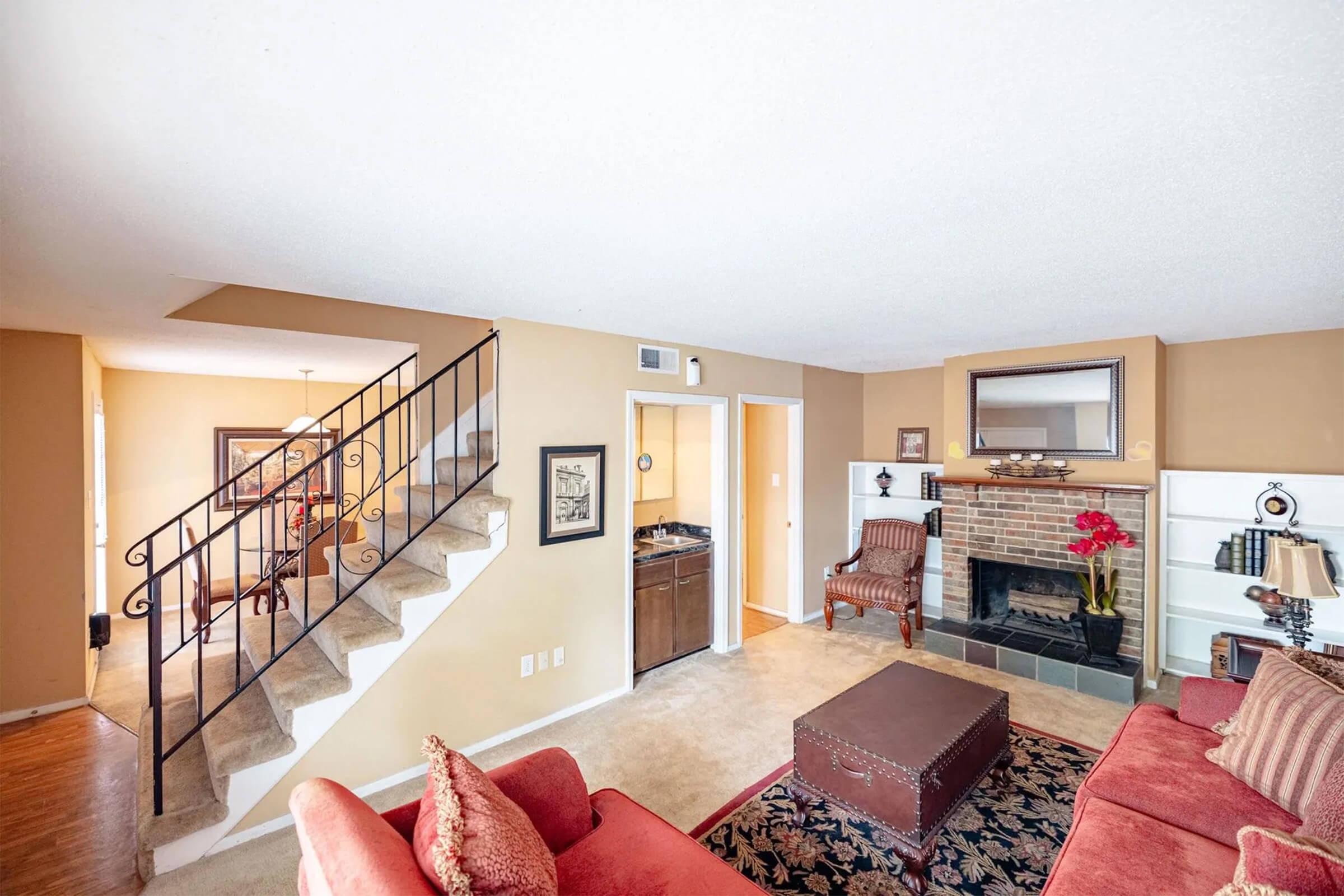 A cozy living room featuring a plush red sofa, a decorative coffee table, and a fireplace with a mirror above it. Stairs lead to an upper level, while a doorway opens to a dining area in the background. The walls are painted a warm beige, and floral decor adds a touch of color.
