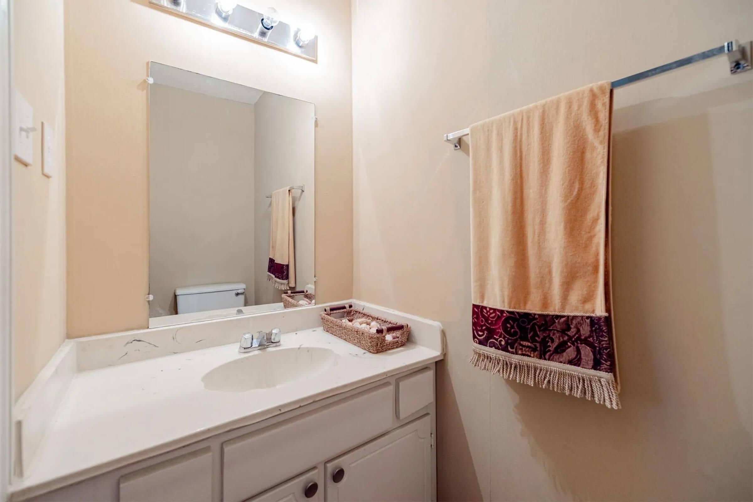 A small bathroom featuring a countertop with a sink, a mirror above, a beige wall color, and a towel hanging on a rack. A basket is placed on the counter, and a toilet is visible in the reflection of the mirror. The decor is minimal and functional.