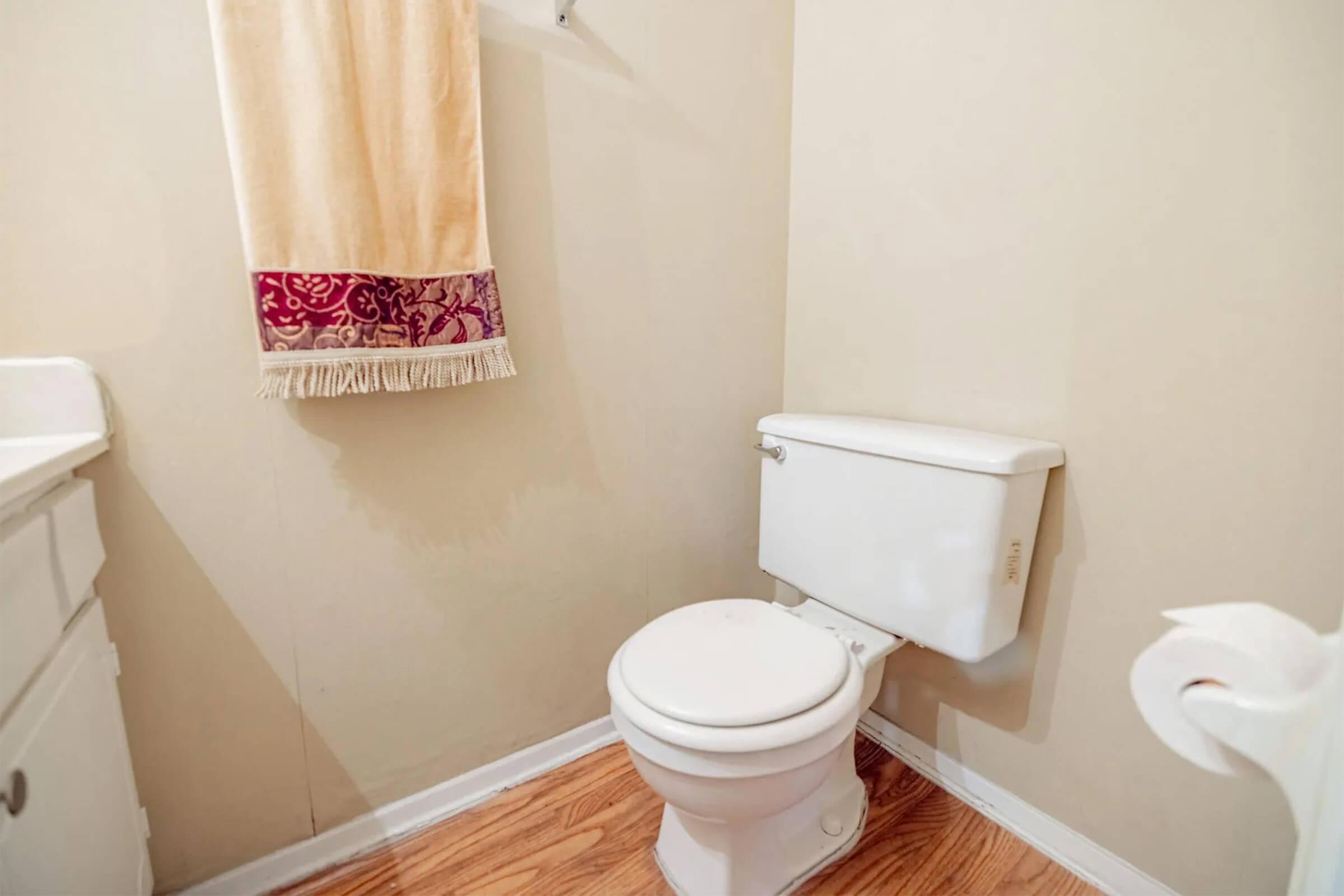 A small bathroom featuring a white toilet, light-colored walls, and a beige towel hanging on the wall with a decorative border. The flooring is wooden, and there is a minimalistic and clean appearance overall.