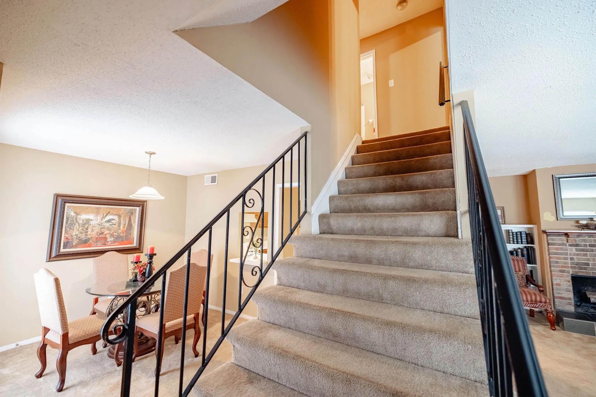 A carpeted staircase leads to an upper level in a well-lit hallway. To the left, a small dining area features a round table with chairs, and a framed artwork on the wall. The walls are painted in warm tones, creating a welcoming atmosphere in the space.