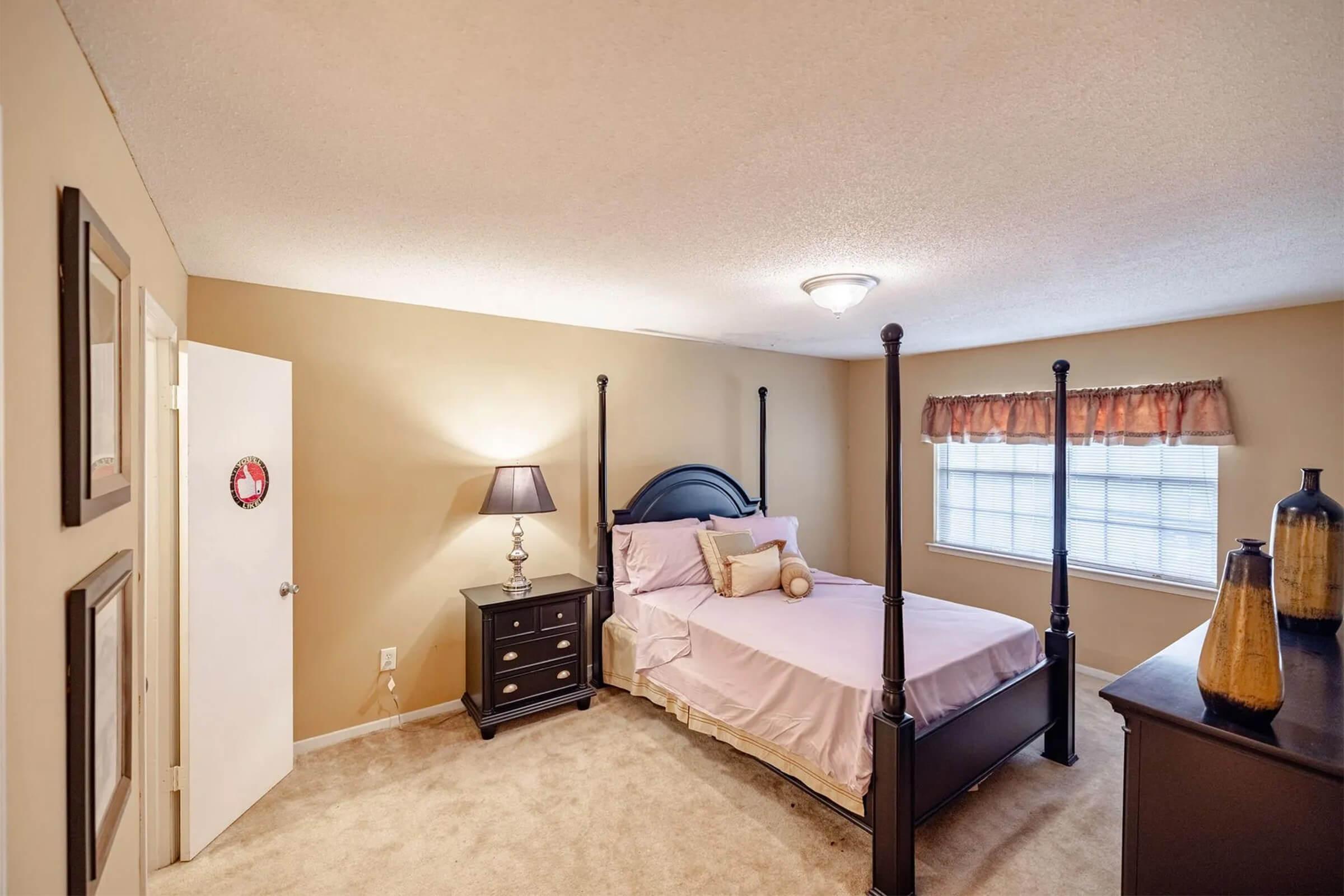 A cozy bedroom featuring a four-poster bed with light bedding, a bedside lamp, and a dark wood nightstand. There is a decorative window treatment and light-colored walls, along with a dresser displaying vases. Soft carpeting enhances the warm, inviting atmosphere of the room.
