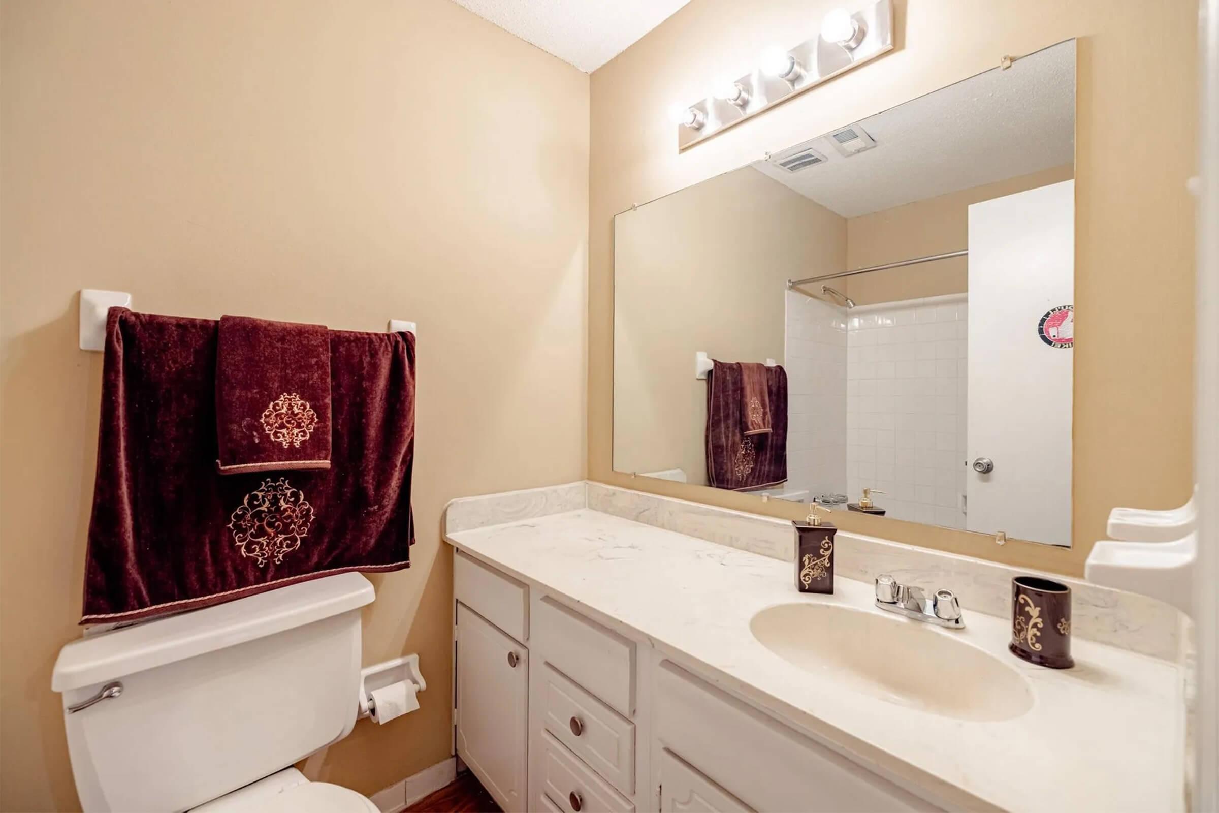 A small bathroom featuring a beige wall color, a white toilet, and a sink with a marble countertop. A large mirror is mounted above the sink, and there are two dark red towels hanging on a rack. The bathroom includes a shower area with a glass door in the background.