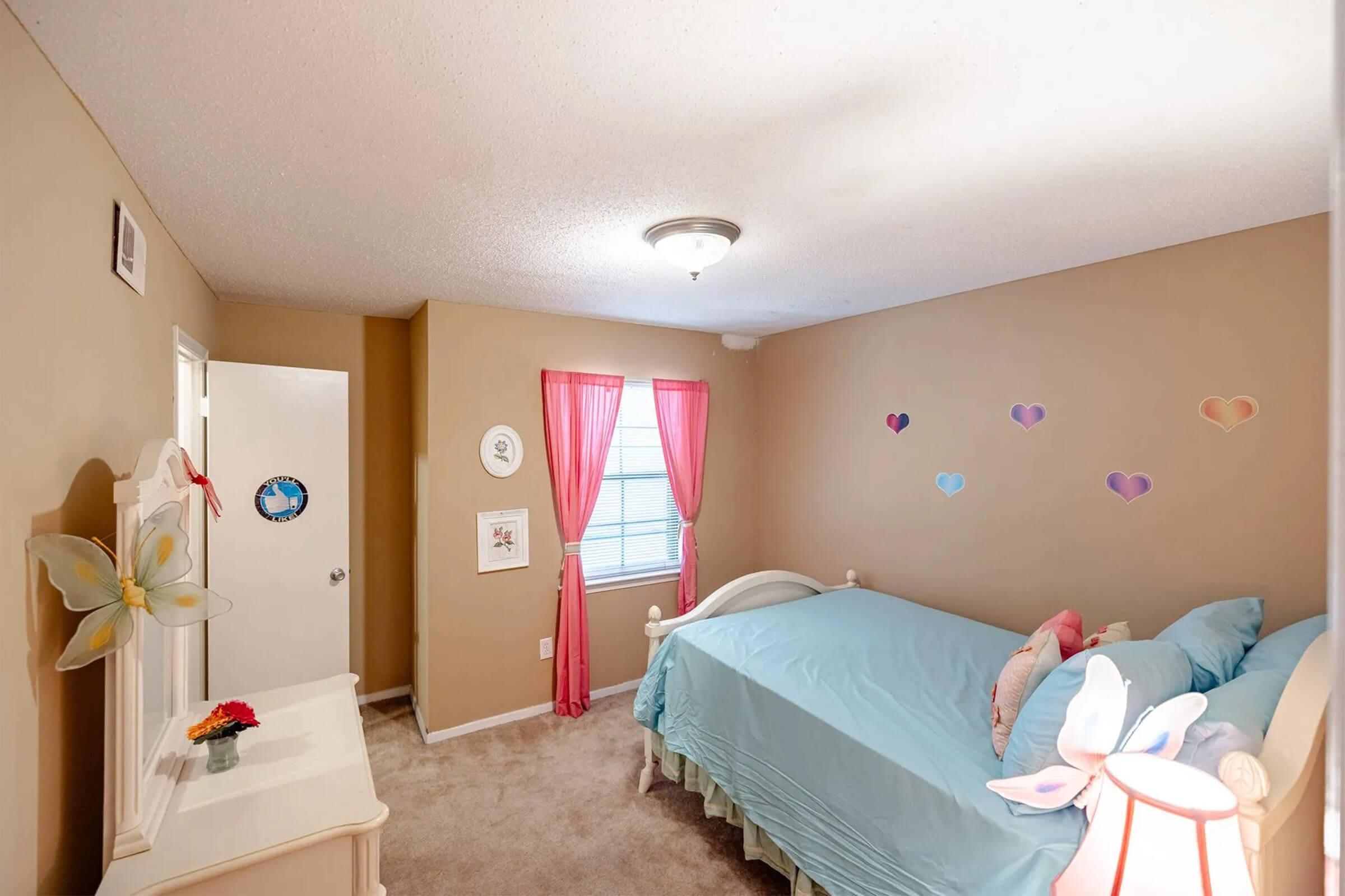 A cozy children's bedroom featuring a light blue bed with decorative pillows, soft beige walls, and a plush carpet. Pink curtains frame a window, and heart-shaped wall decals add colorful touches. A white dresser and a small flower vase enhance the inviting atmosphere.