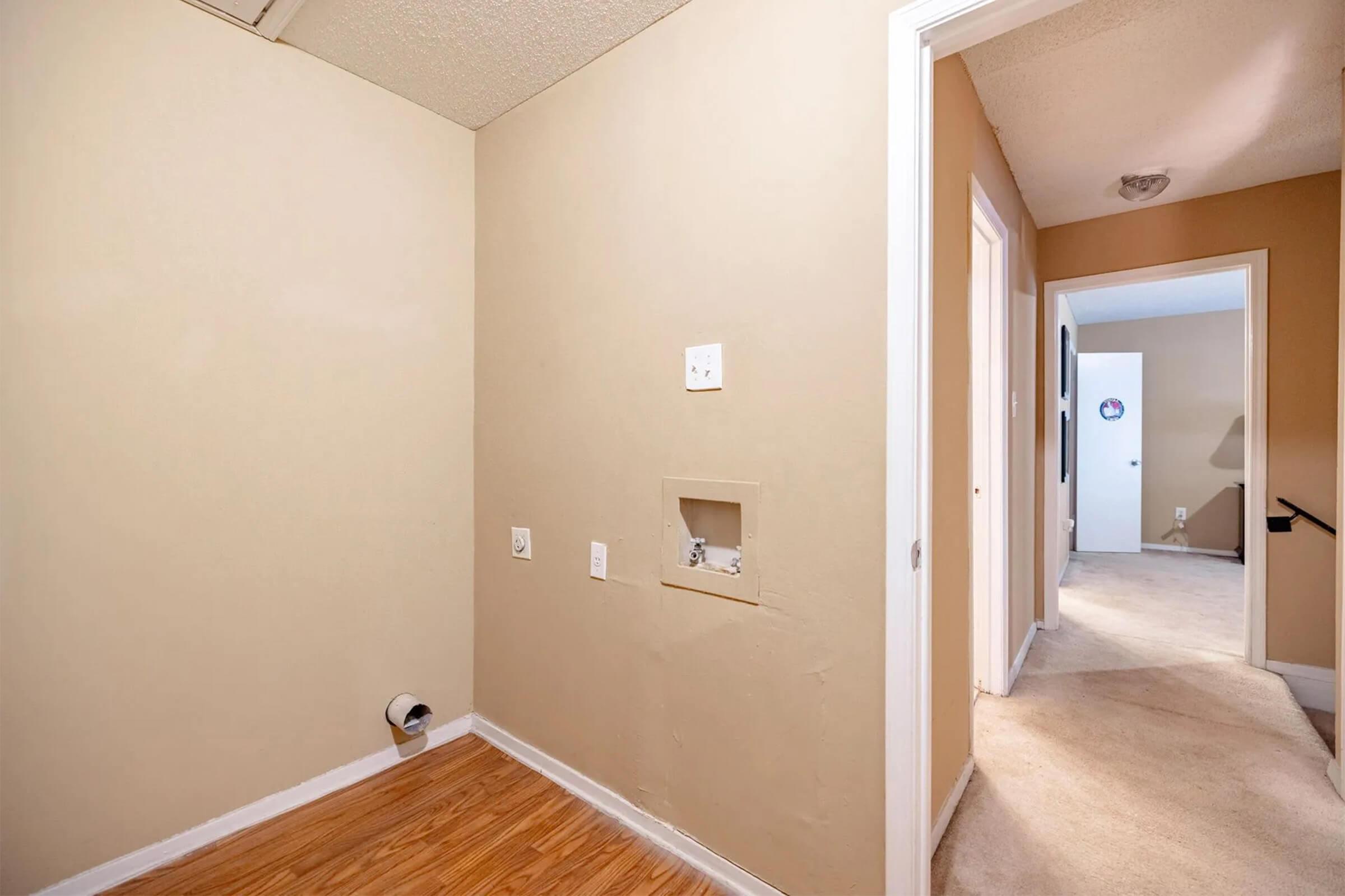 Narrow hallway with light beige walls and wooden flooring. On the left, there's an empty space with a power outlet and a small recessed area. A door frame can be seen on the right leading to another room. Soft lighting and carpeted flooring are visible in the background.