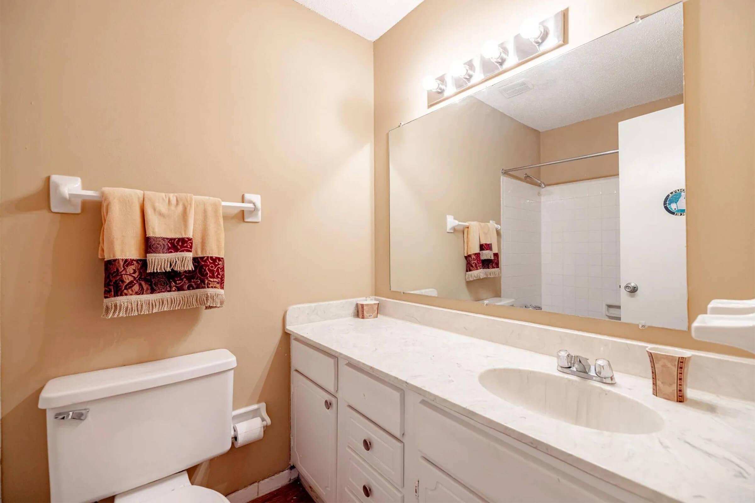 A bathroom with beige walls, a white marble countertop, a large mirror, a towel rack with a decorative towel, and a simple sink. The toilet is positioned next to a cabinet with drawers, and a shower area is visible behind a glass door at the back. Soft lighting is provided by overhead fixtures.
