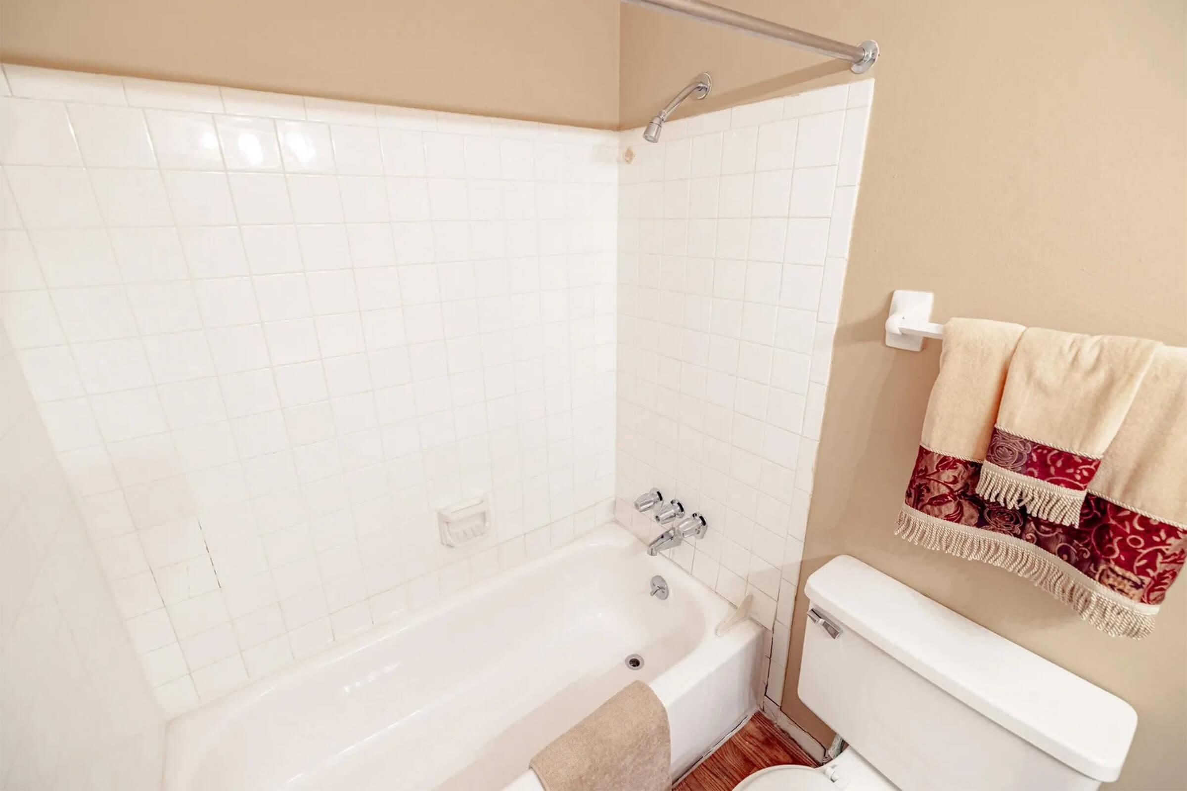 A clean bathroom featuring a white bathtub with a shower, a white tiled wall, a toilet, and a towel rack with a decorative towel. The floor is wooden, creating a warm contrast with the white fixtures.