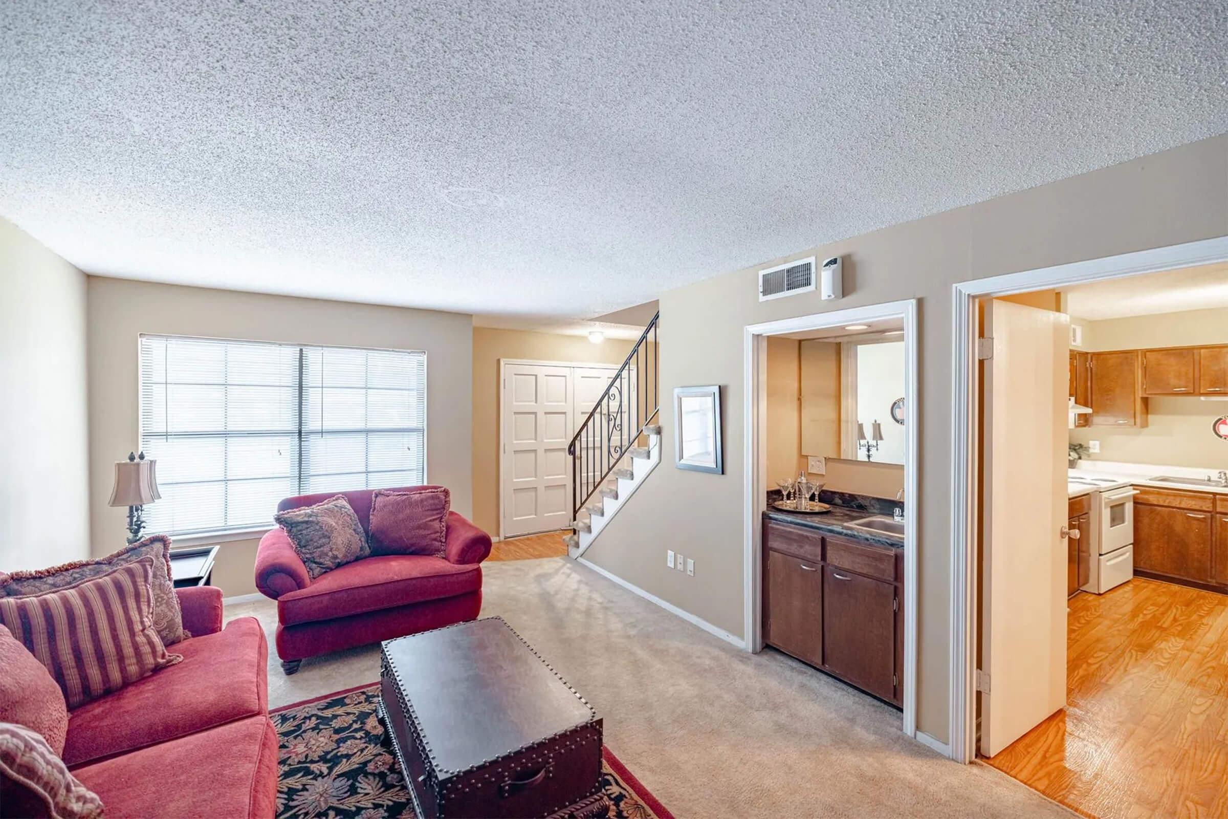 A cozy living room featuring a red sofa and armchair, a coffee table, and a staircase leading to the upper level. There are large windows allowing natural light, and an open kitchen can be seen through an adjoining doorway. The interior has light-colored walls and neutral carpeting.