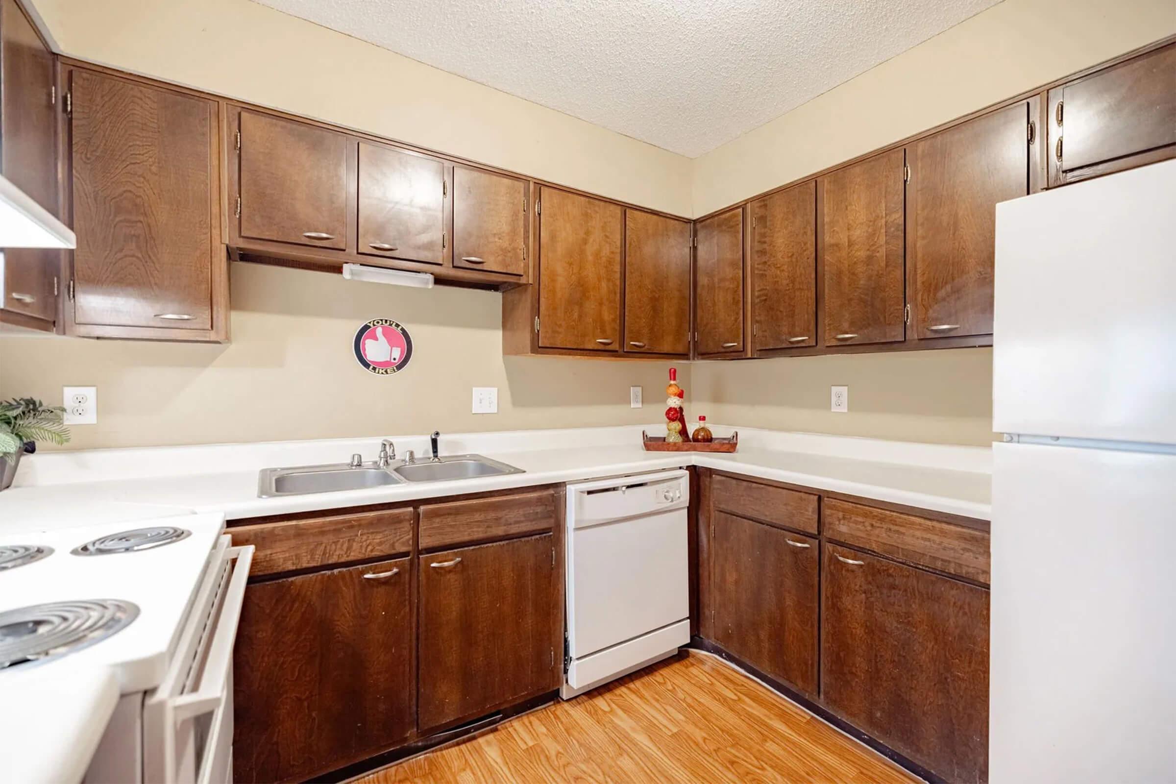 A well-lit kitchen featuring dark wooden cabinets, a white refrigerator, and a sink with a dishwasher. The countertops are light-colored, and there are decorative items on display, including a round wall clock and a few kitchen utensils. The flooring is a warm wood laminate.