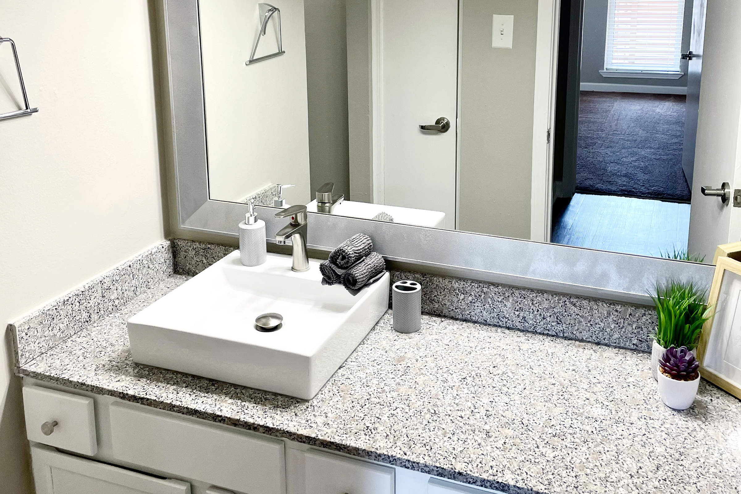A modern bathroom sink with a white square basin on a granite countertop. Neatly arranged are gray towels, a soap dispenser, and a toothbrush holder. A mirror reflects the space, and a small potted plant adds a touch of greenery. The background shows a doorway leading to a well-lit room.
