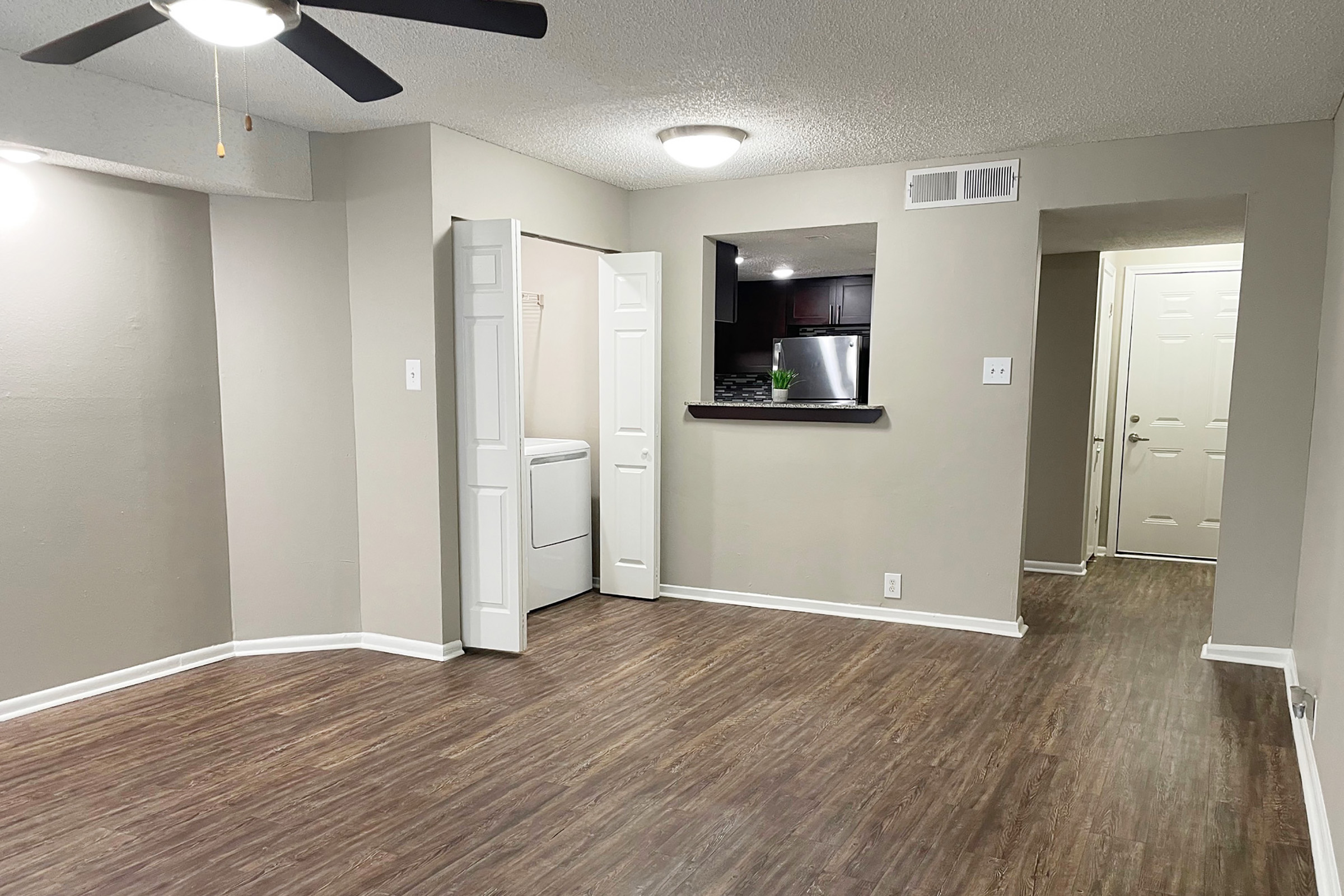 Interior view of a modern apartment featuring a spacious living area with wooden flooring, a ceiling fan, and neutral-colored walls. There is a doorway leading to a laundry area, a breakfast bar open to a kitchen space with dark cabinets, and an entrance visible on the right. Soft overhead lighting enhances the ambiance.