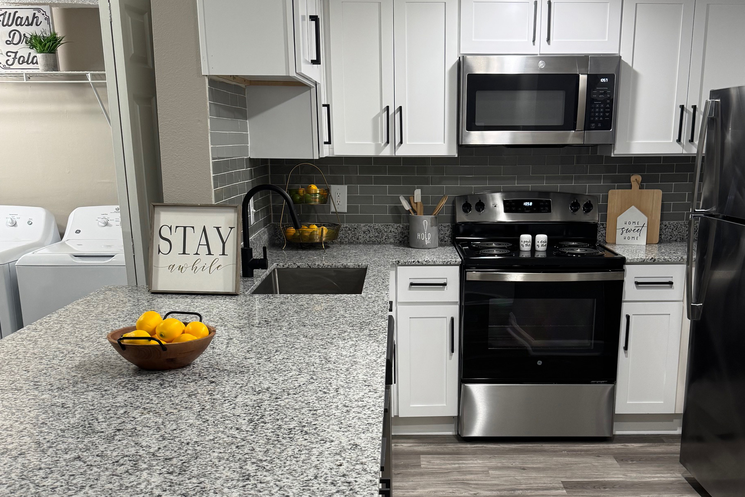 Modern kitchen featuring gray granite countertops, white cabinets, and stainless steel appliances. A bowl of lemons sits on the counter, with a small decorative sign that says "STAY AWHILE." In the background, there are laundry appliances and a dish drying rack, creating a cozy and inviting space.