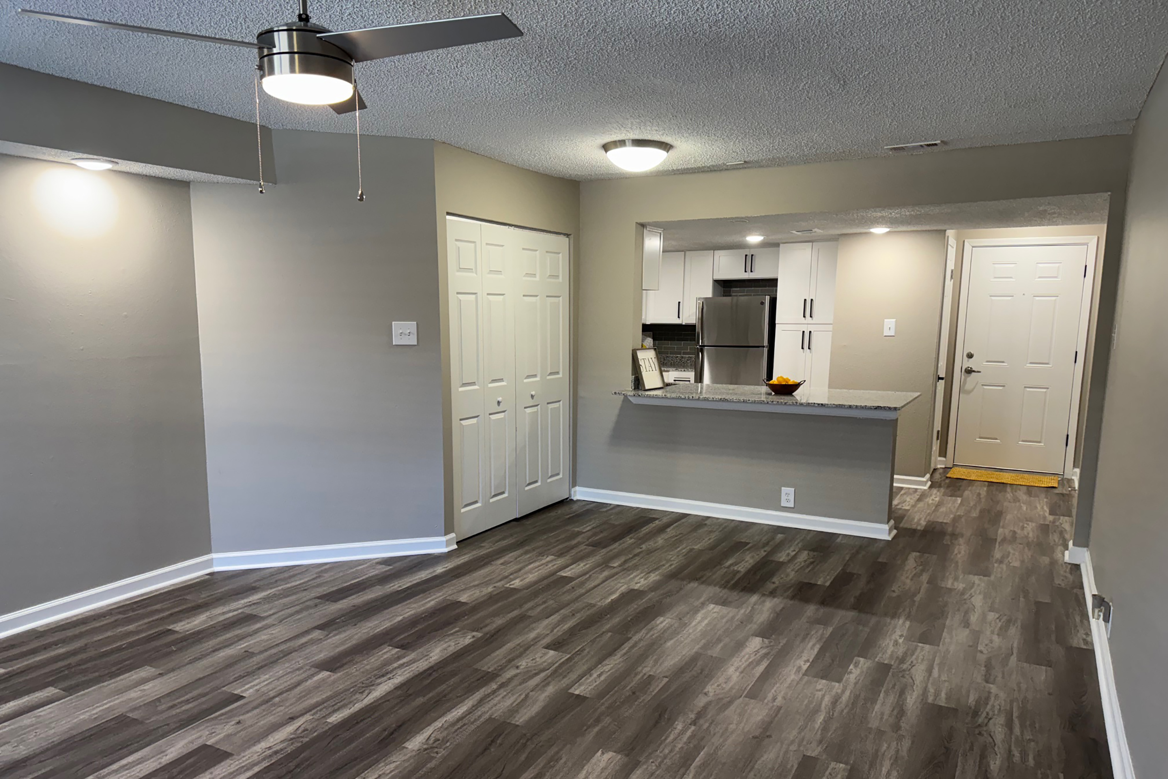 Interior view of an apartment showcasing a spacious living area with light gray walls and laminate flooring. A ceiling fan is visible, and there are two white closet doors. In the background, a modern kitchen area can be seen with a granite countertop and stainless steel appliances. A doorway leads outside.