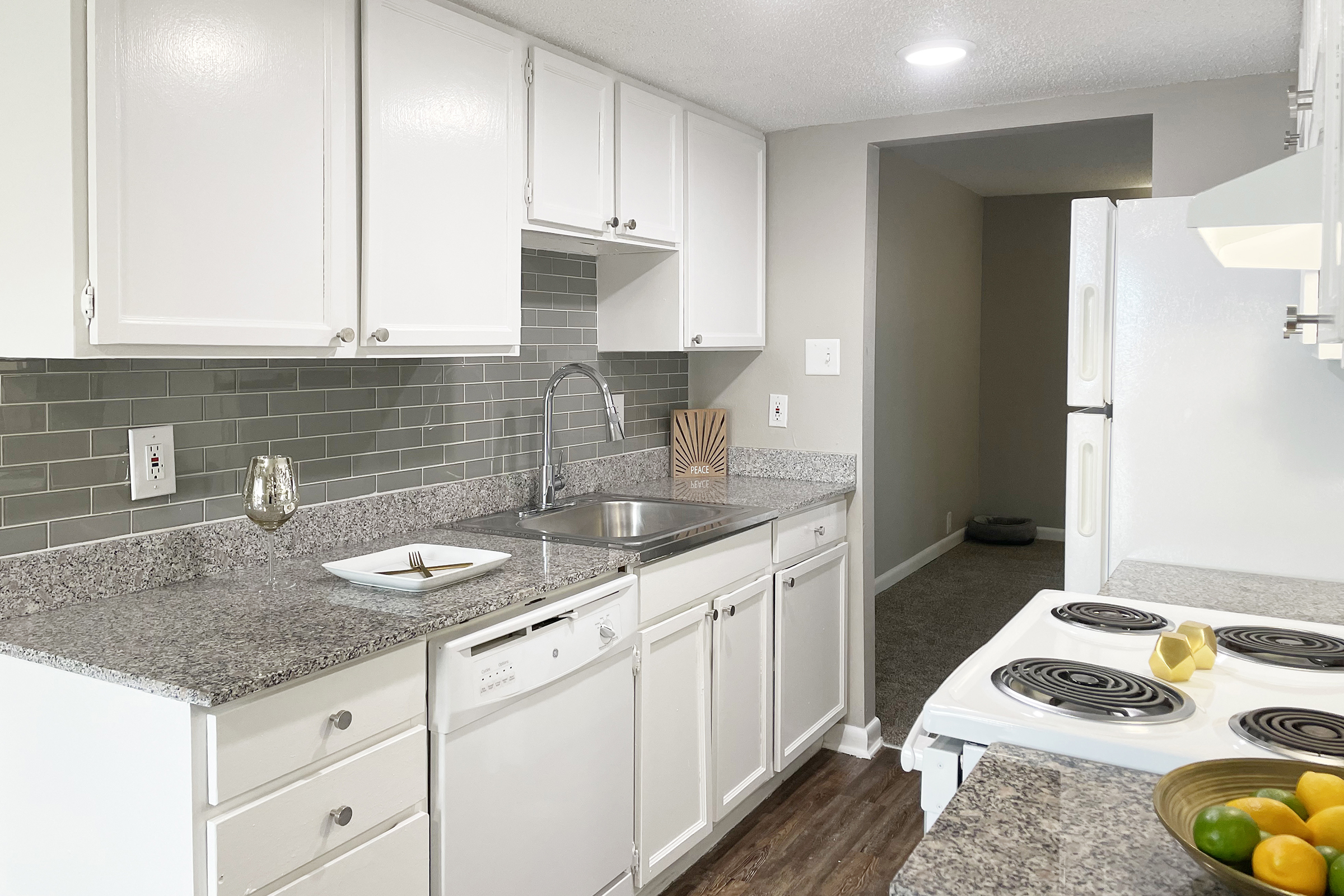 A modern kitchen featuring white cabinetry, gray tiled backsplash, and granite countertops. The sink is to the left with a decorative item nearby. The stove is situated on the right, and the room has a light-colored wall with an open doorway leading to another space. A glass of wine and a plate are on the countertop.