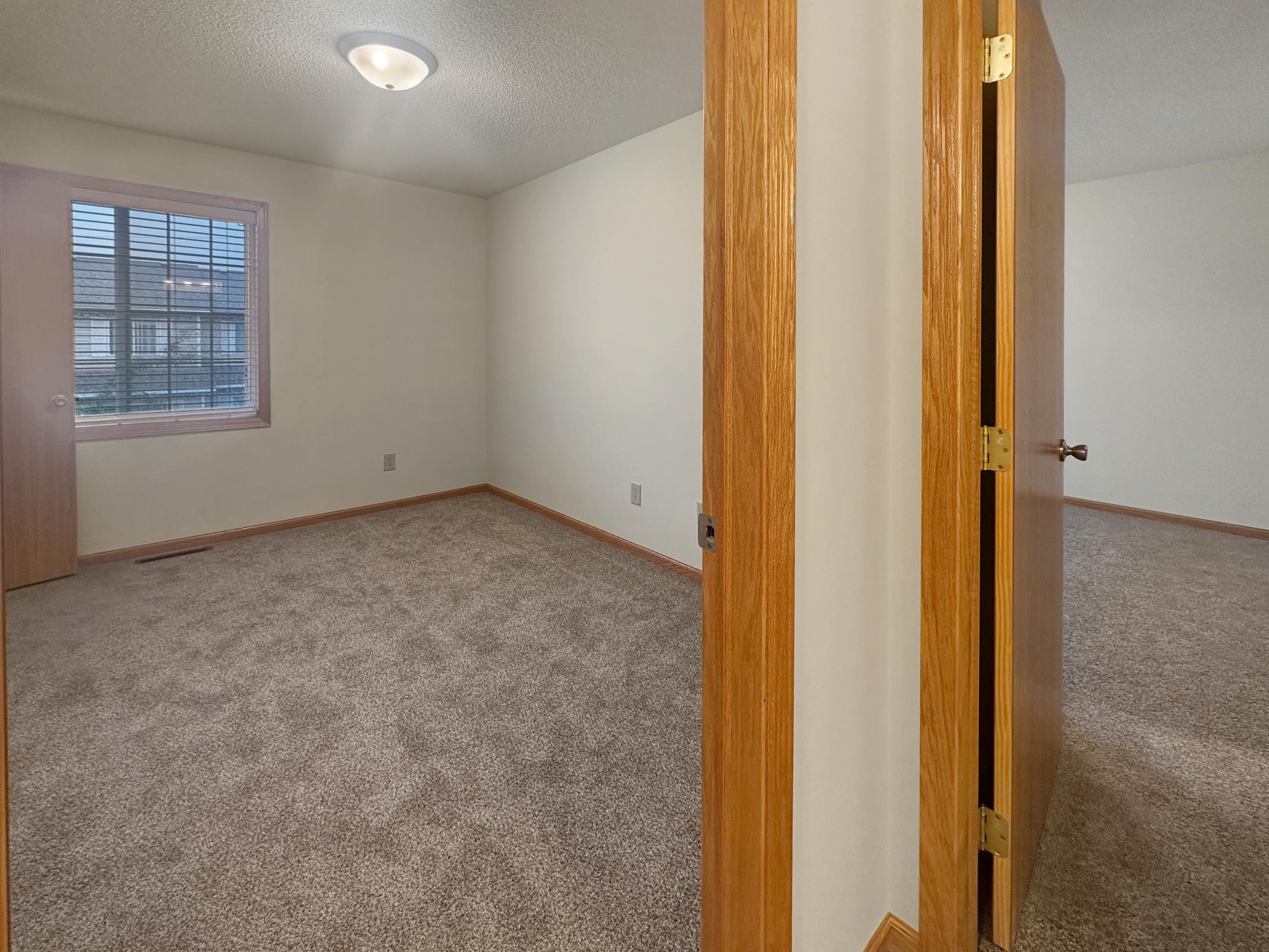 A vacant room with light beige walls and a textured carpet. The doorway is visible on the right, leading to another part of the space. Natural light enters through a window on the left, which has blinds. The room appears well-lit and clean, suitable for various uses like a bedroom or office.