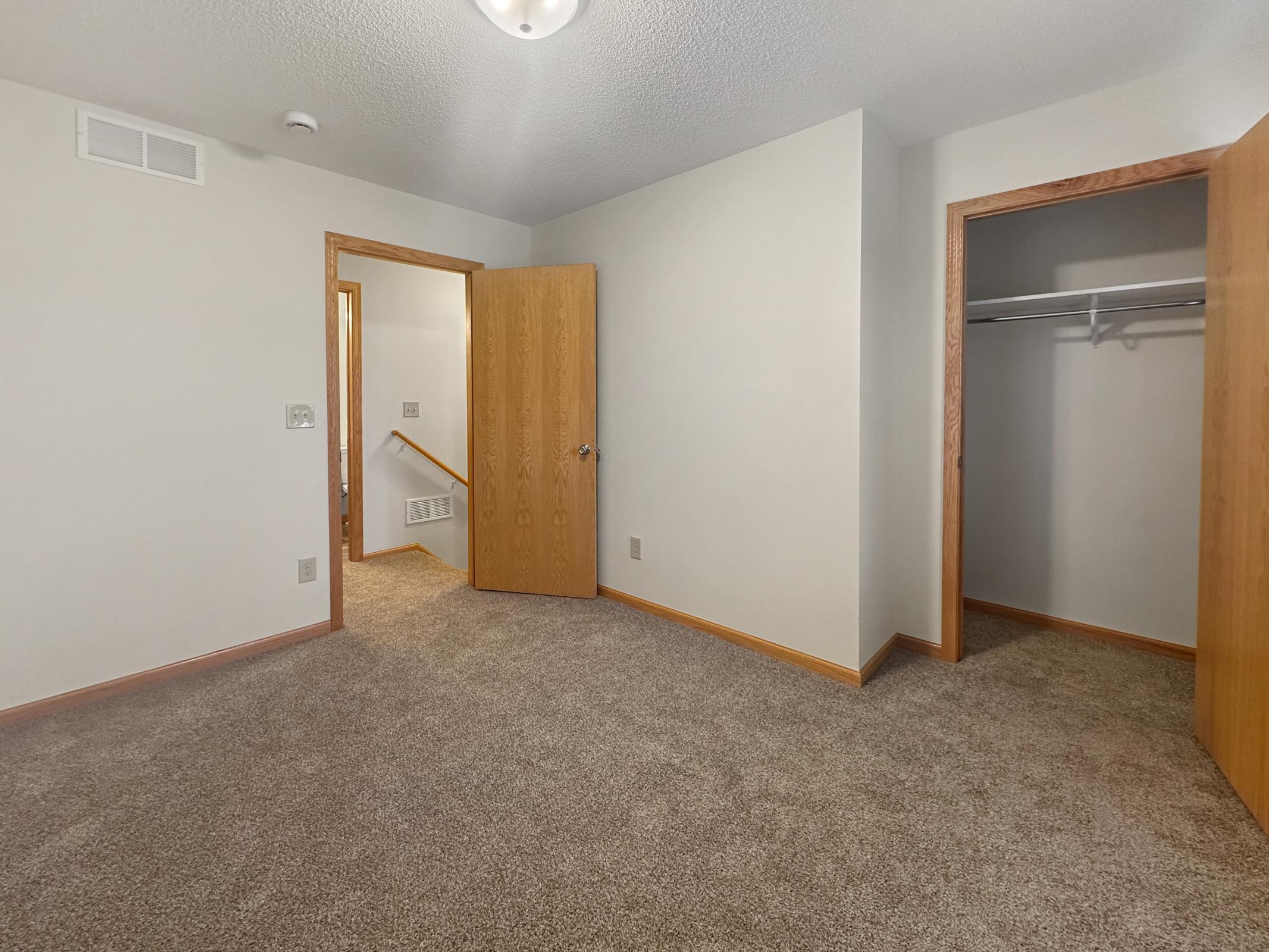 A partially furnished room featuring beige walls and carpet. There are two wooden doors: one leading to a closet and another to a hallway. Natural light filters in, highlighting the space's neutral tones, creating a cozy and inviting atmosphere.