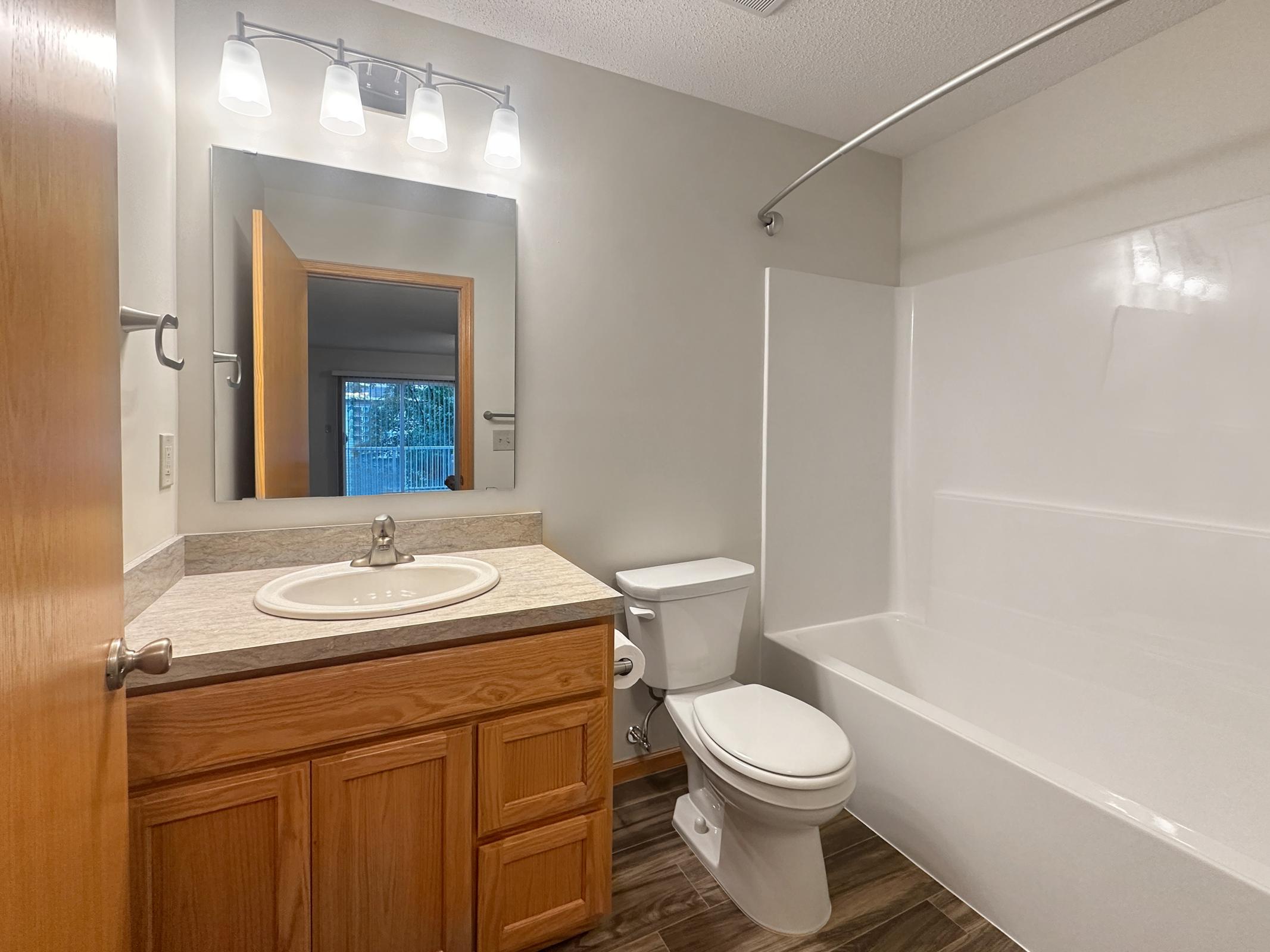 A clean, bright bathroom featuring a white bathtub with a shower, a wooden vanity with a sink, and a toilet. The walls are painted a light color, and there is a large mirror above the sink. Soft lighting from the fixtures adds warmth to the space. A door leads to another room visible in the background.