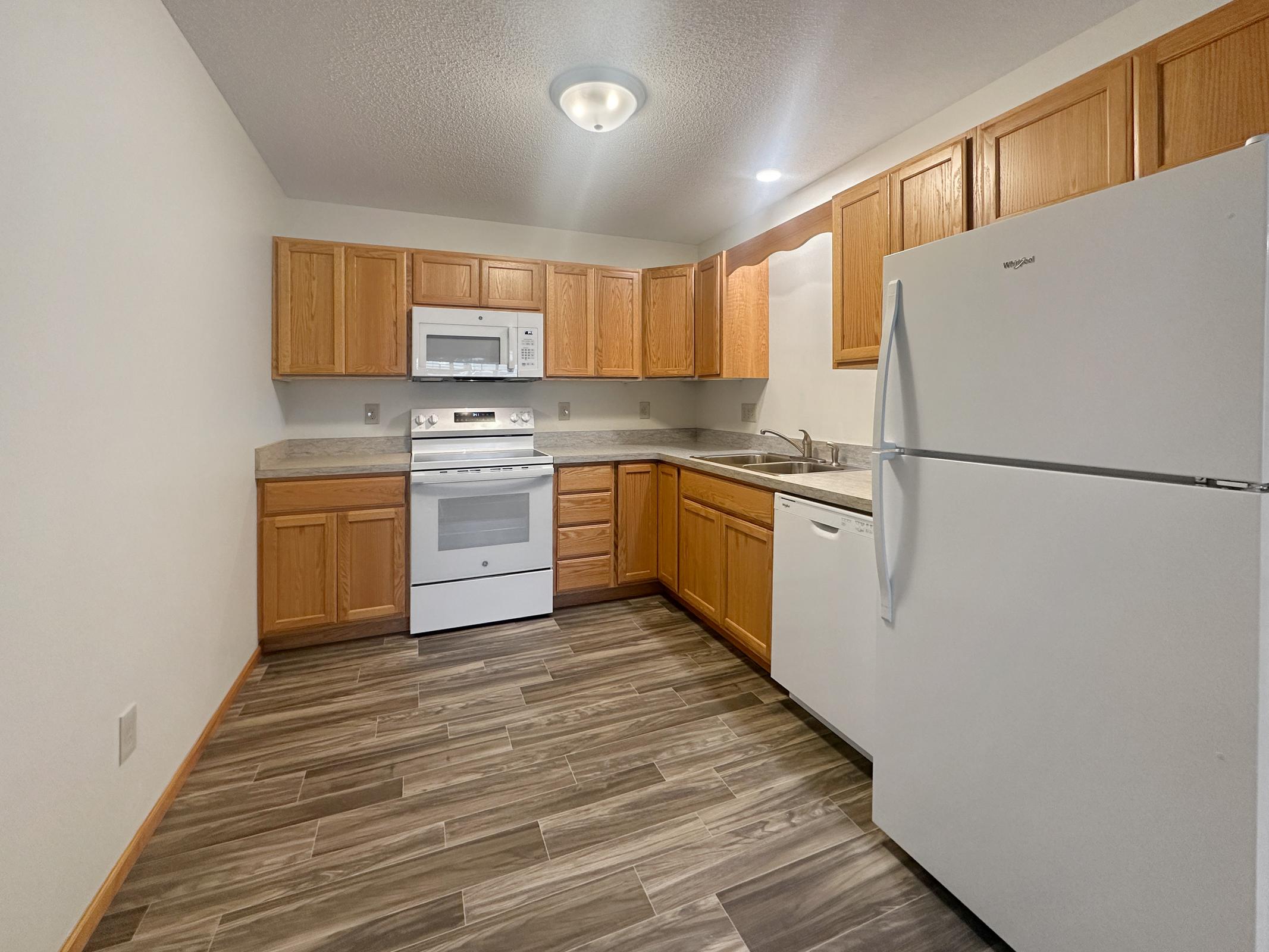 A modern kitchen featuring wooden cabinets, a white refrigerator, a white oven with an overhead microwave, and a dishwasher. The countertop is light-colored with a speckled design, and the floor is tiled in a dark, wood-like pattern. Soft lighting illuminates the space.